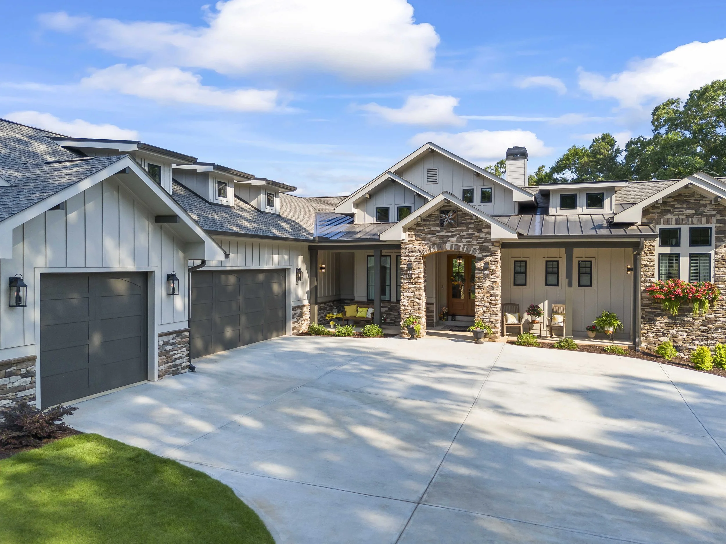 Front view of a modern house with a driveway, green lawn, and a porch with chairs and potted plants. The house has white siding, stone accents, multiple gabled roofs, and a chimney. The sky is blue with some clouds.