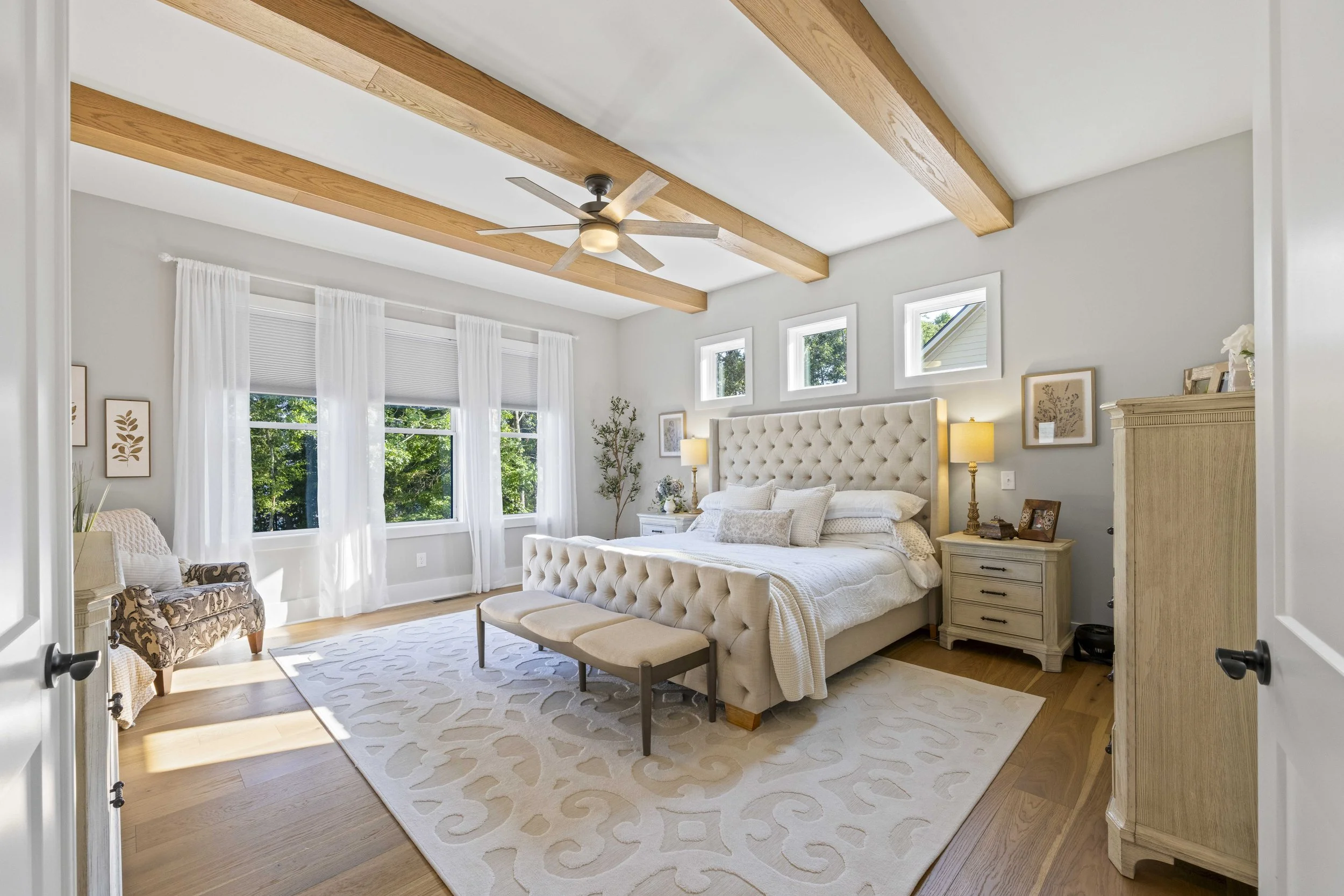 A bright bedroom with a white tufted bed, nightstands with lamps, large windows with white curtains, and a ceiling fan with wooden beams.