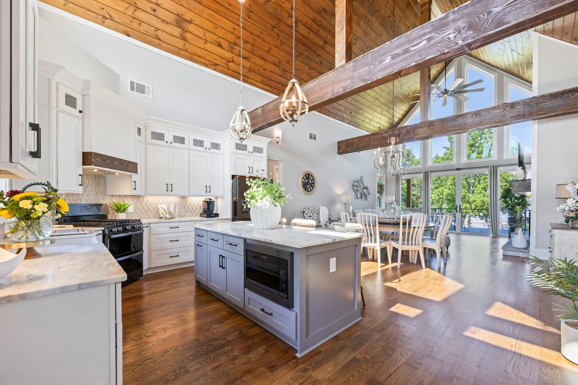 Open-concept kitchen and dining area with vaulted wood ceiling, pendant lights, and large windows showing trees outside.