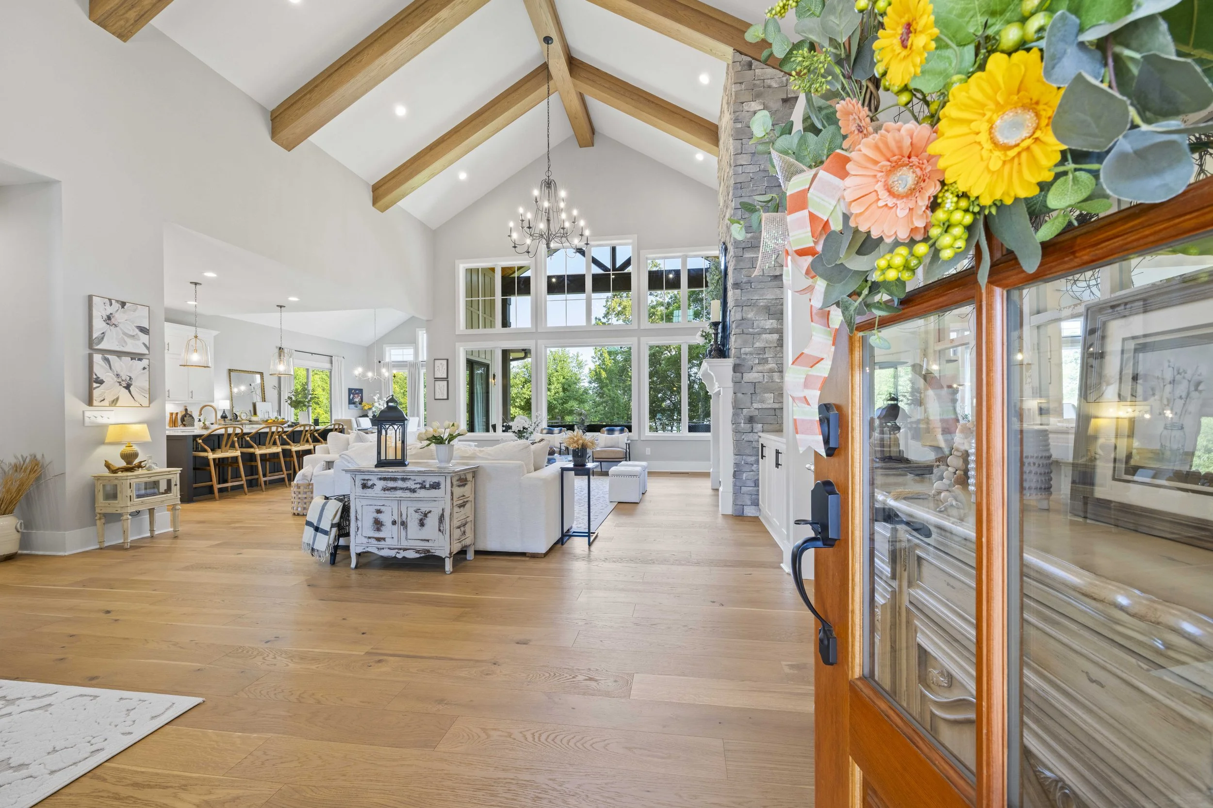 Open living room with high vaulted ceiling, exposed wooden beams, large windows, a stone fireplace, and various furniture including a white sofa, coffee table, and dining area in the background. There are colorful flowers and a wooden display cabinet