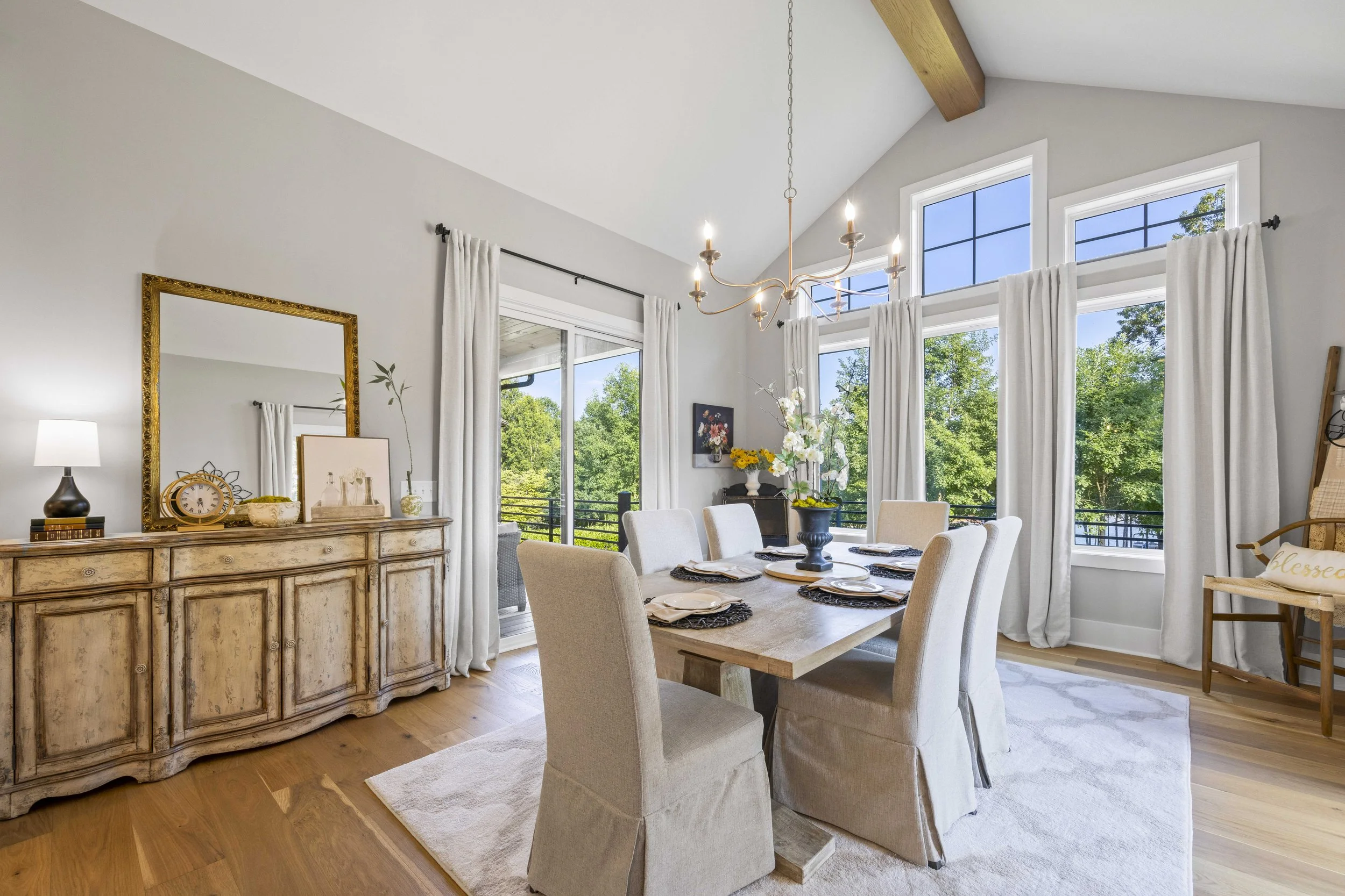Bright dining room with a rustic wooden sideboard, large mirror, white curtains, a chandelier, and a table set with plates and napkins near large windows showing green trees outside.