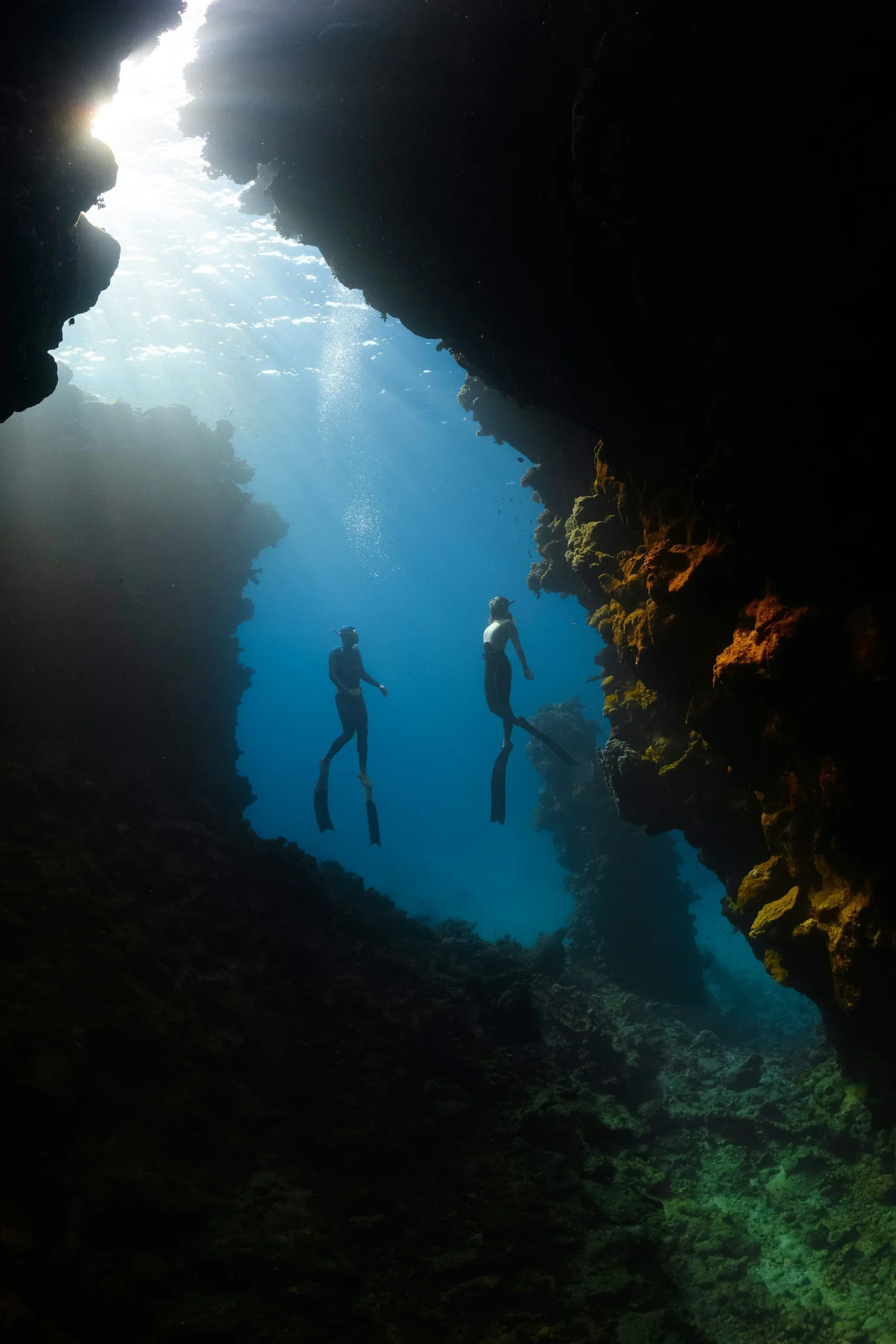 Divers silhouetted against blue water beyond an underwater rock opening