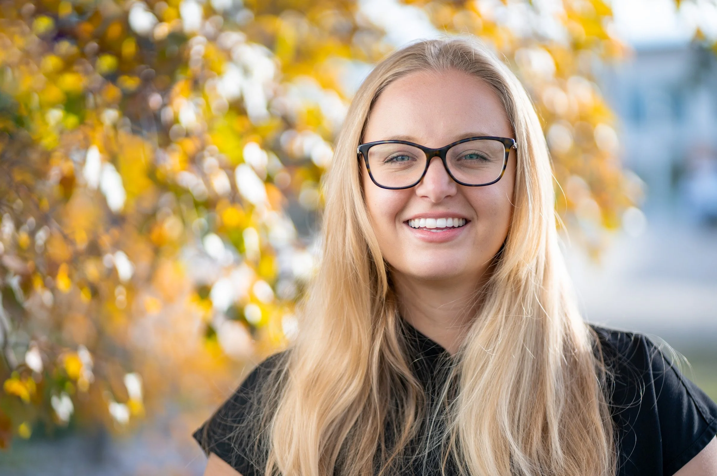 A young woman with long blonde hair, wearing glasses and a black top, smiling outdoors with fall foliage in the background.