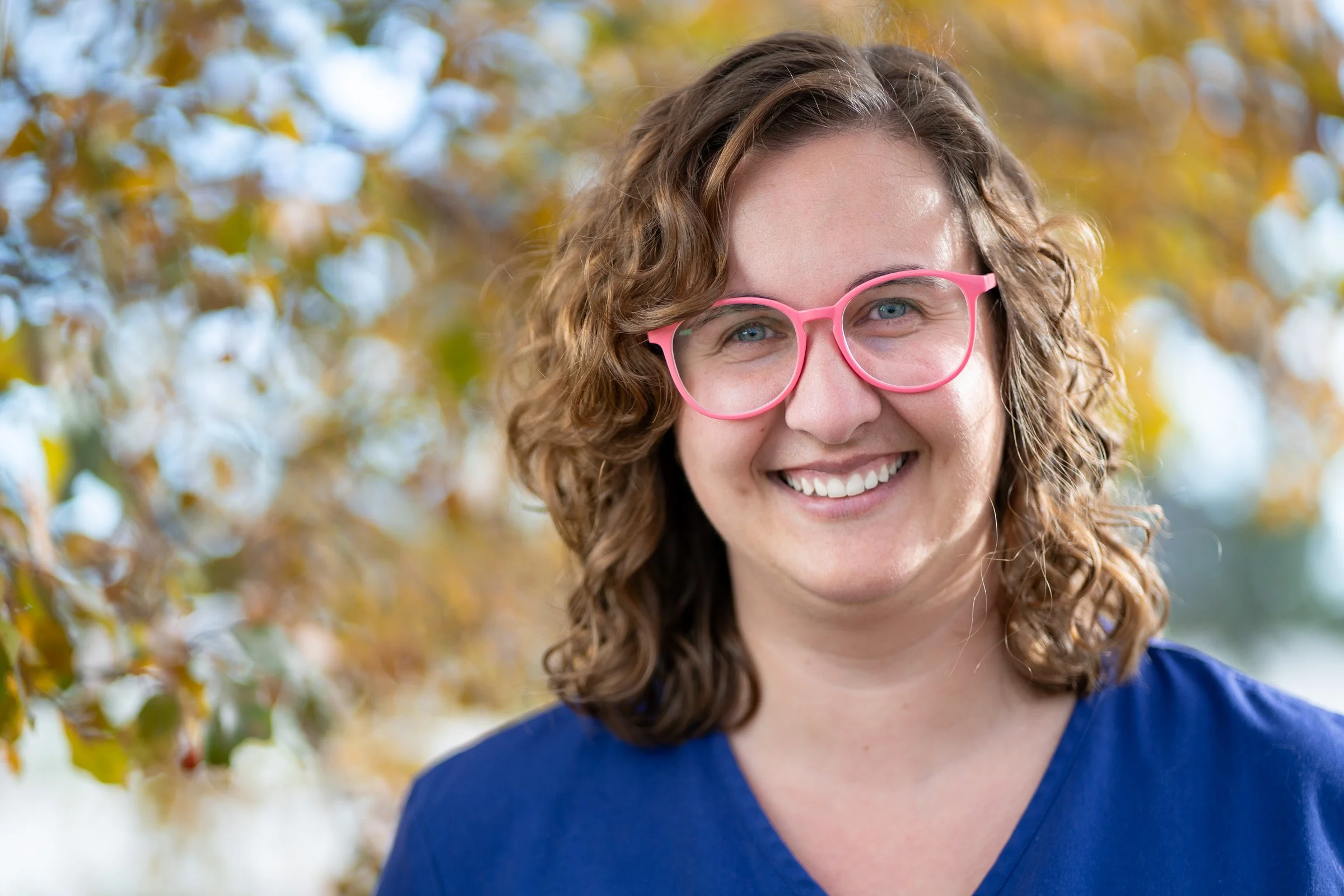 A smiling woman with curly brown hair wearing pink glasses and a blue shirt outdoors with fall-colored leaves in the background.