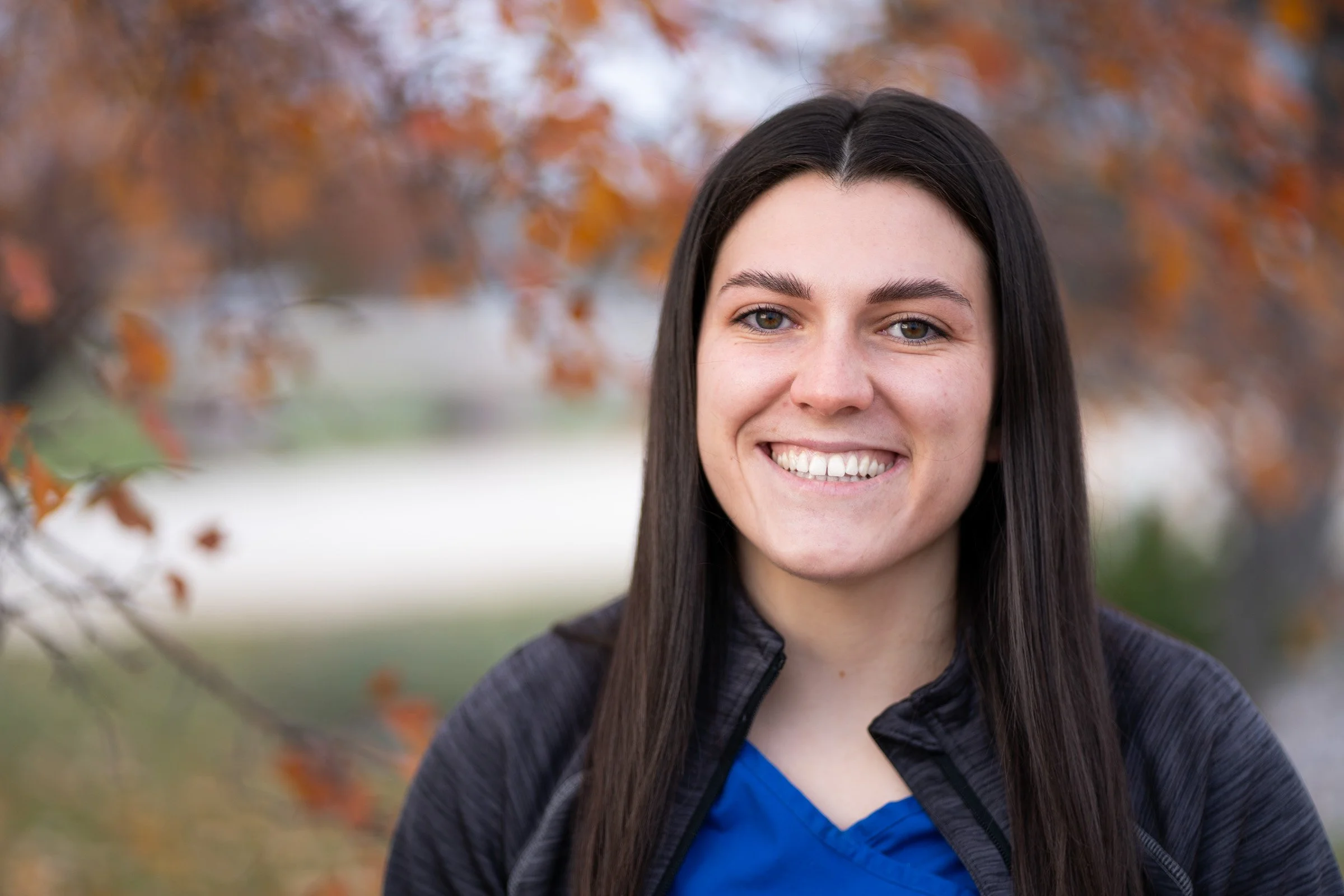 A young woman with long dark hair, smiling, standing outdoors with autumn-colored trees in the background.
