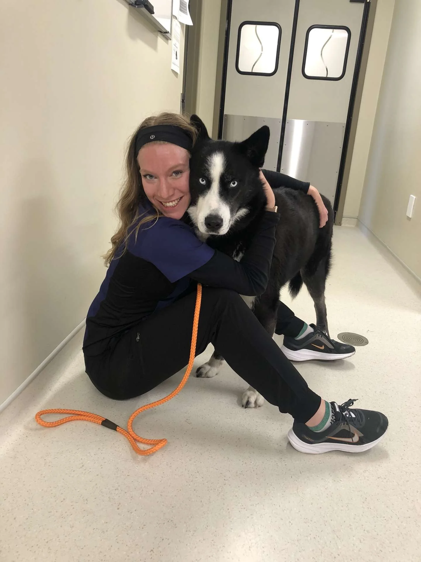 A woman in athletic clothing hugging a black and white Siberian Husky dog while sitting on the floor in a hallway near a door.