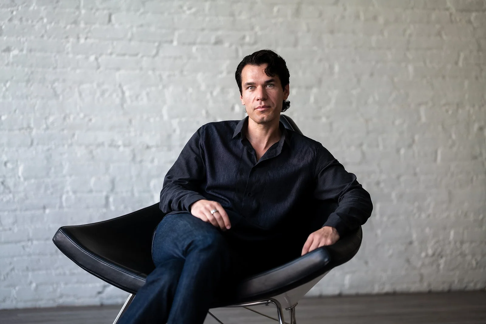 A man with dark hair, wearing a black shirt and blue jeans, sitting on a modern black chair in front of a white brick wall.