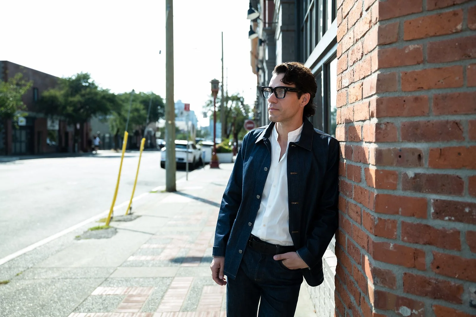 A man with dark hair and glasses leaning against a red brick wall on a city sidewalk, wearing a white shirt and dark jacket, looking off into the distance on a sunny day.