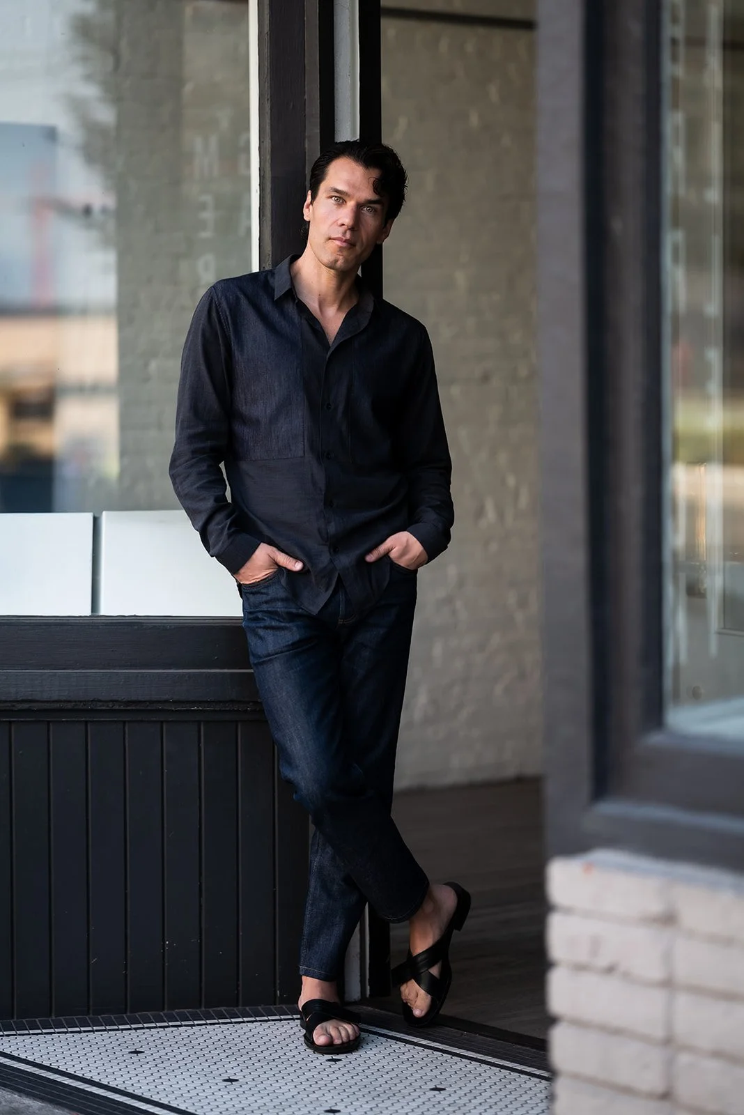 A man standing outside a building next to a window, wearing a black shirt, jeans, and black sandals, with hands in pockets and leaning against the wall.