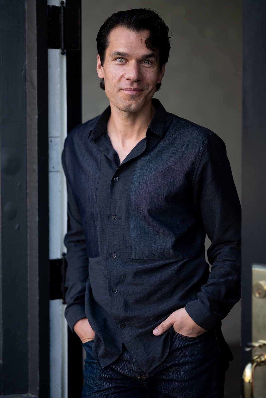 A man with dark hair and light skin wearing a black button-up shirt, standing by a doorway with one hand in his pocket, smiling at the camera.