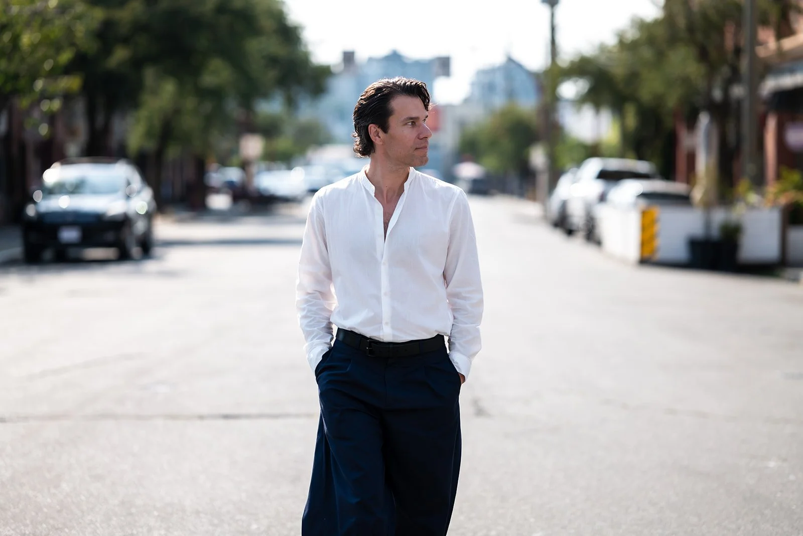 A man in a white shirt and navy pants walking on an urban street with parked cars and trees in the background.