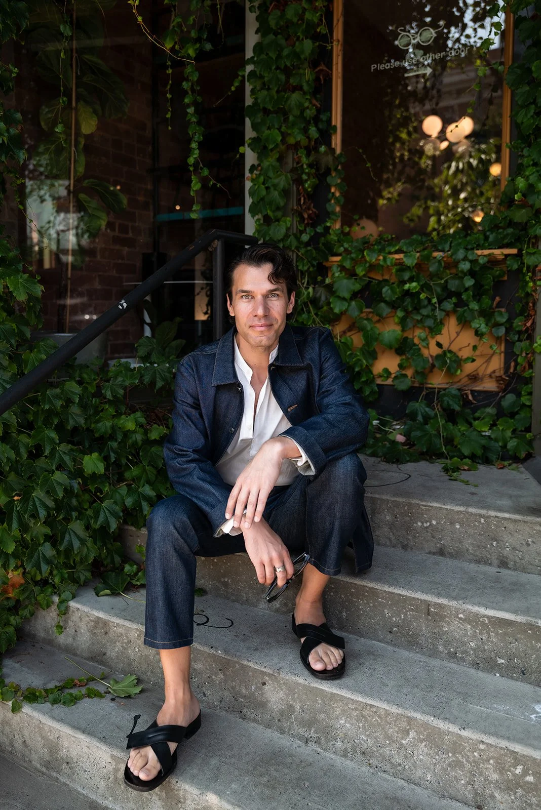 A man with dark hair, wearing a denim jacket, white shirt, dark jeans, and black sandals, sitting on outdoor stone steps surrounded by green ivy and plants.