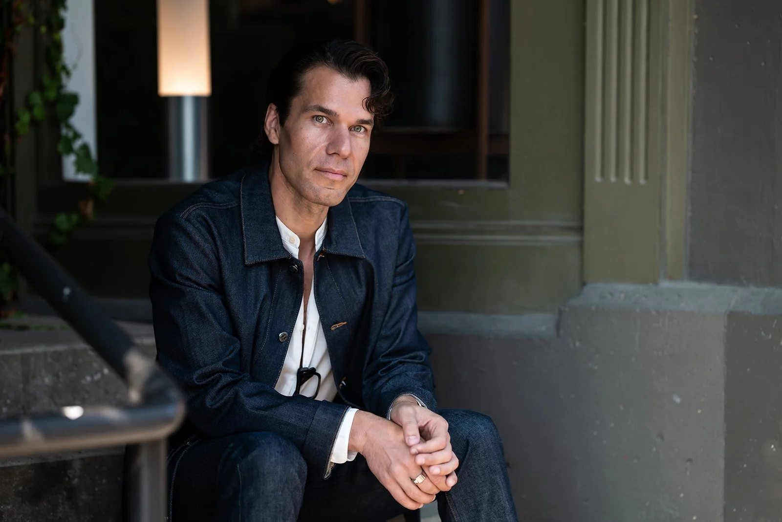 A man with dark hair and light eyes sitting on a stoop outside a building, wearing a denim jacket and a white shirt, with a neutral expression.