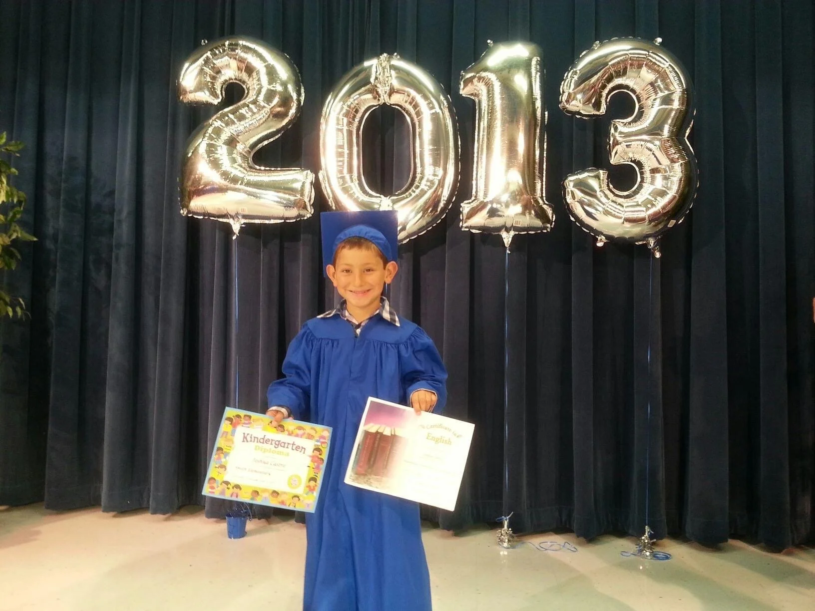 A young boy dressed in a blue graduation gown and cap standing in front of large silver balloons spelling out 2013. He is holding two certificates/papers, one with the title 'Kindergarten Diploma'.