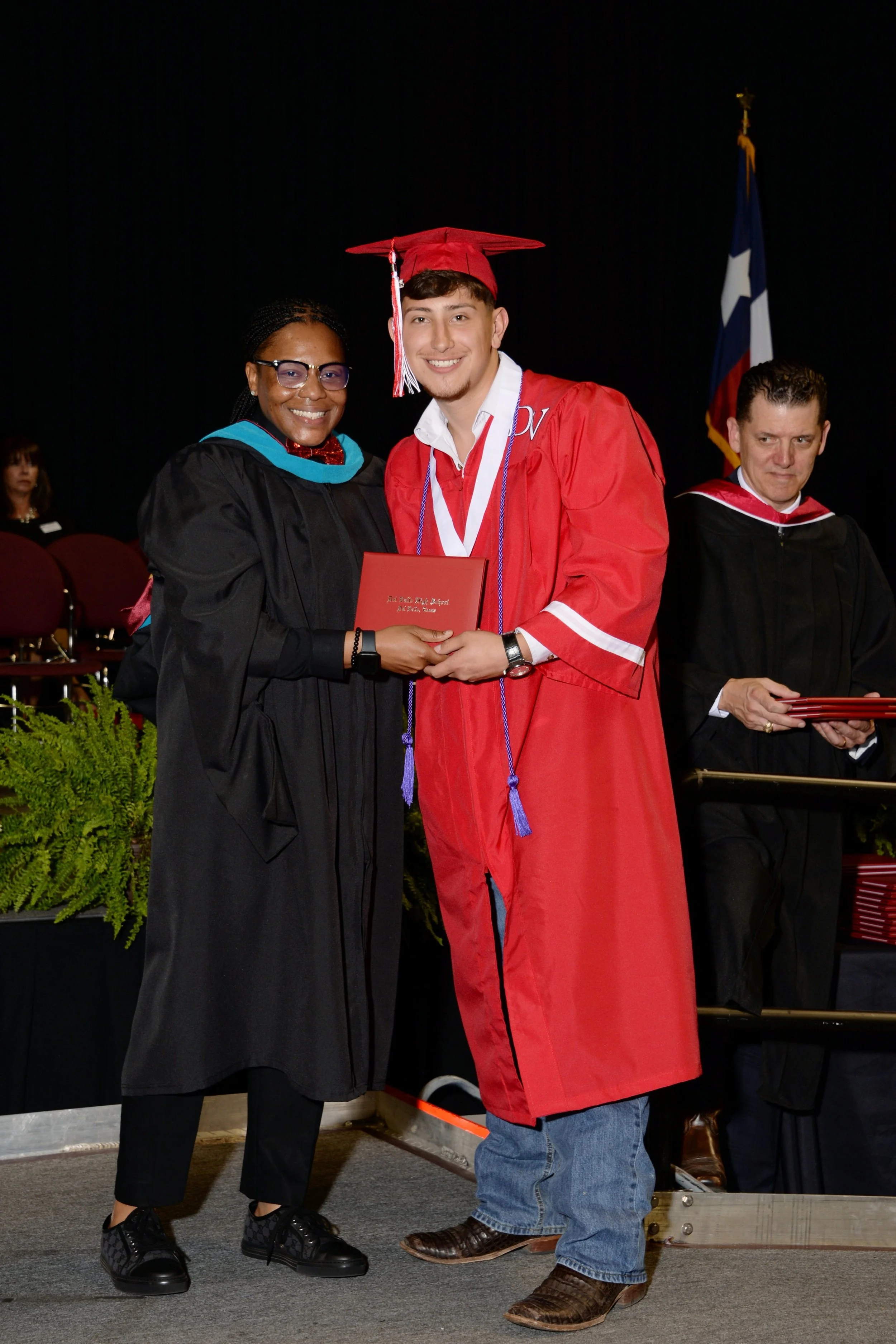 A young man in a red graduation gown and cap receiving his diploma from a woman in black academic regalia at a graduation ceremony. Another man in black academic regalia stands behind them. The background is dark with a flag visible on the right side.