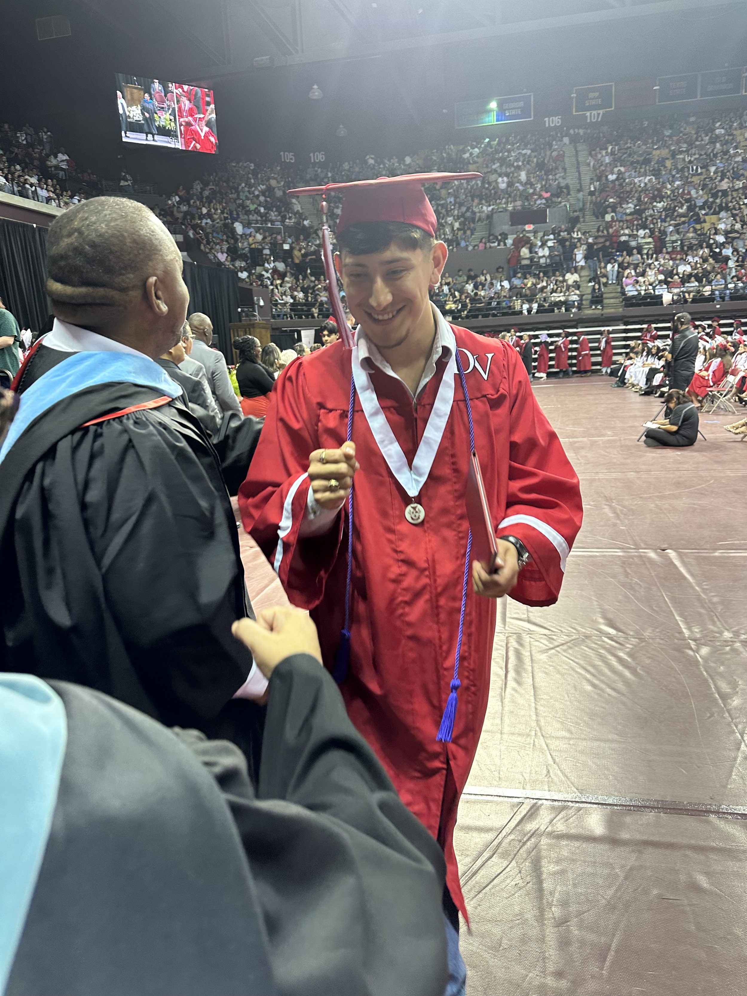 A young man in a red graduation gown and cap smiling as he shakes hands with an older man in academic regalia at a graduation ceremony inside a packed stadium. Graduated from Del Valle High School
