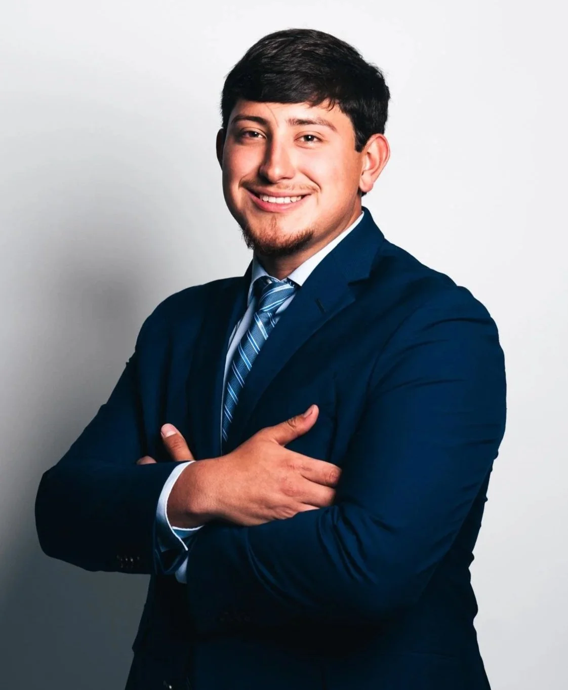 A young man dressed in a dark blue suit with a matching striped tie, smiling, with arms crossed in front of a plain white background.
