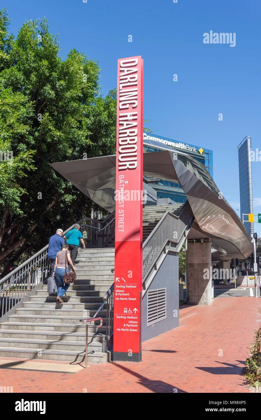 People walking up outdoor stairs near a large red sign that reads "Darling Harbour" and "Bathurst Street" with directions to nearby locations, with modern buildings and trees in the background.