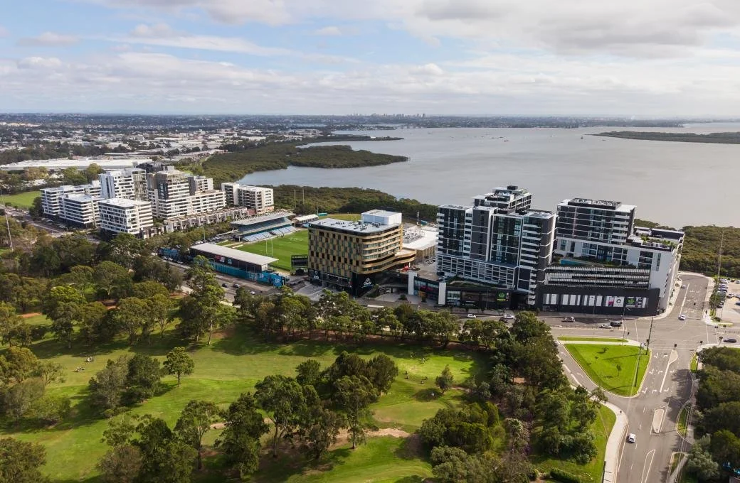 Aerial view of a cityscape with modern buildings near a large body of water, surrounded by green parks and trees.