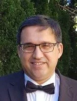 A man wearing glasses, a tuxedo with a bow tie, smiling outdoors with trees in the background.