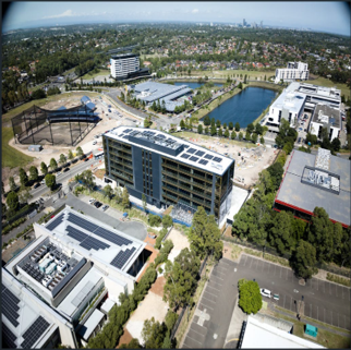 An aerial view of a cityscape showing modern buildings, a large pond, and a sports stadium surrounded by greenery and roads.