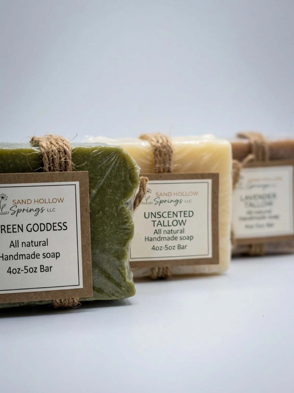 Three handmade soap bars from Sand Hollow Springs LLC, labeled Green Goddess, Unscented Tallow, and a third soap. The soap bars are wrapped with twine and have labels with product details.