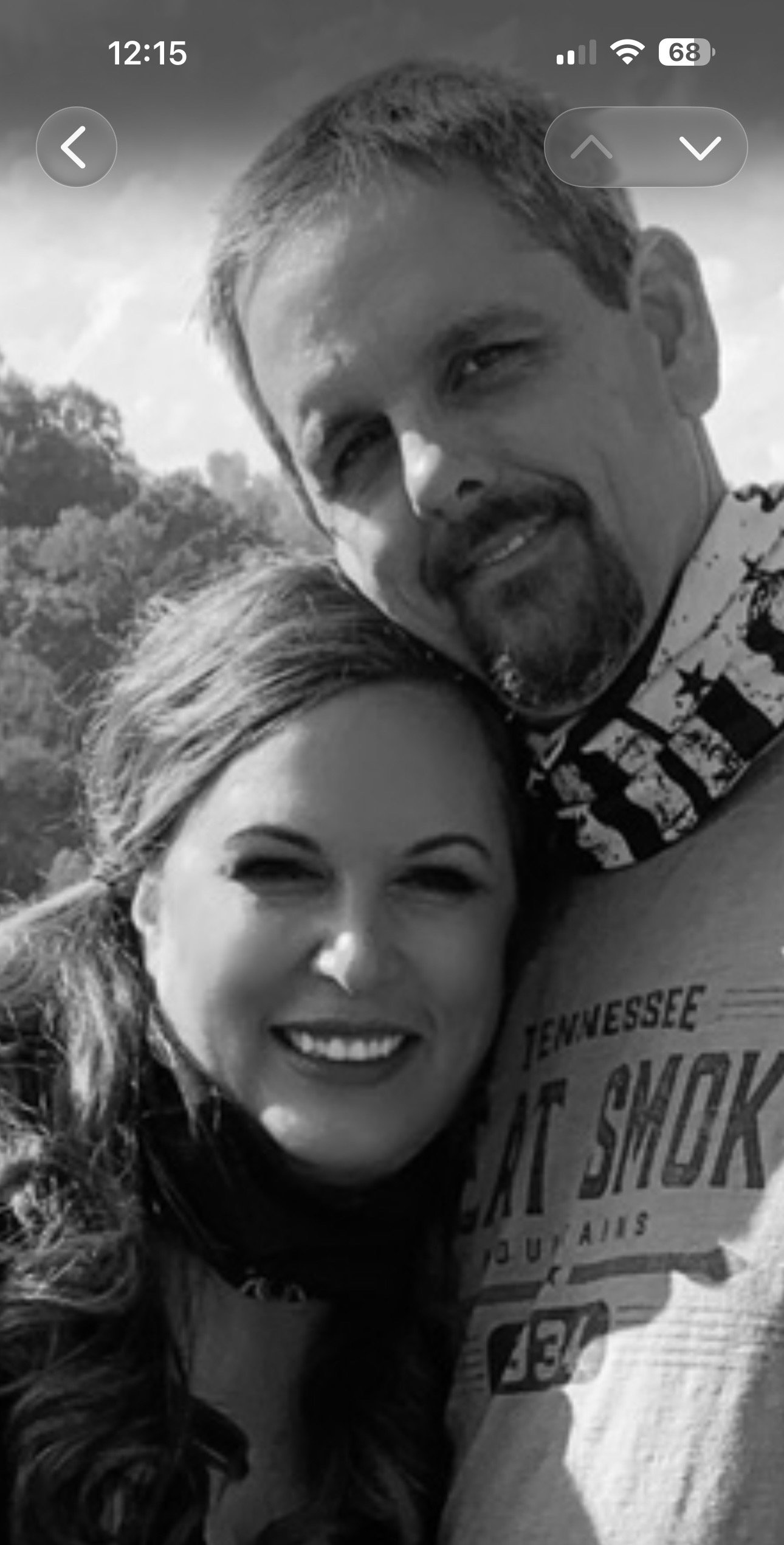 Black and white photo of a smiling woman with long wavy hair and a man with short hair, goatee, and mustache. They are outdoors with trees in the background.