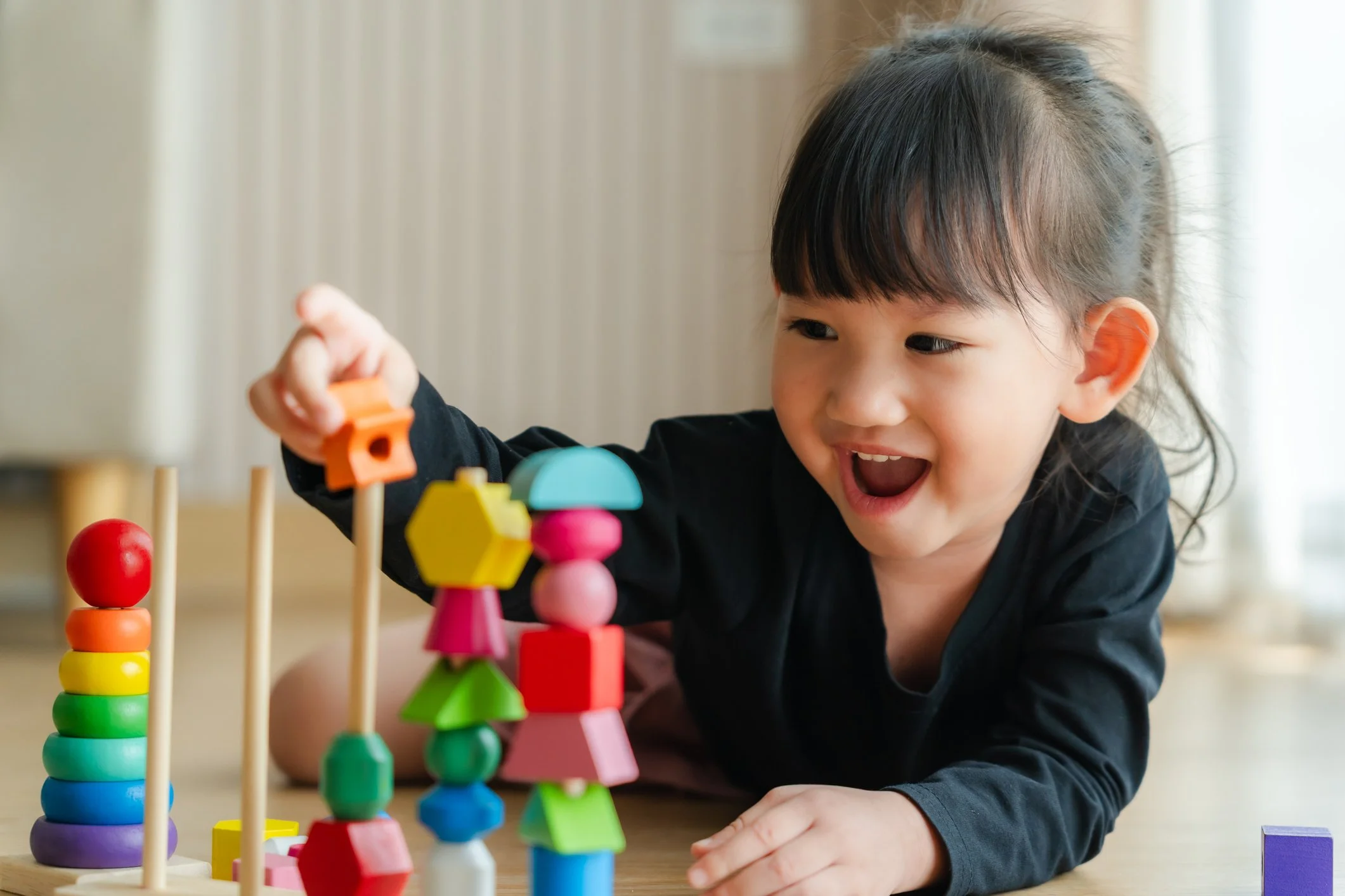 A young girl with dark hair and bangs playing with colorful stacking toys on a wooden surface indoors.
