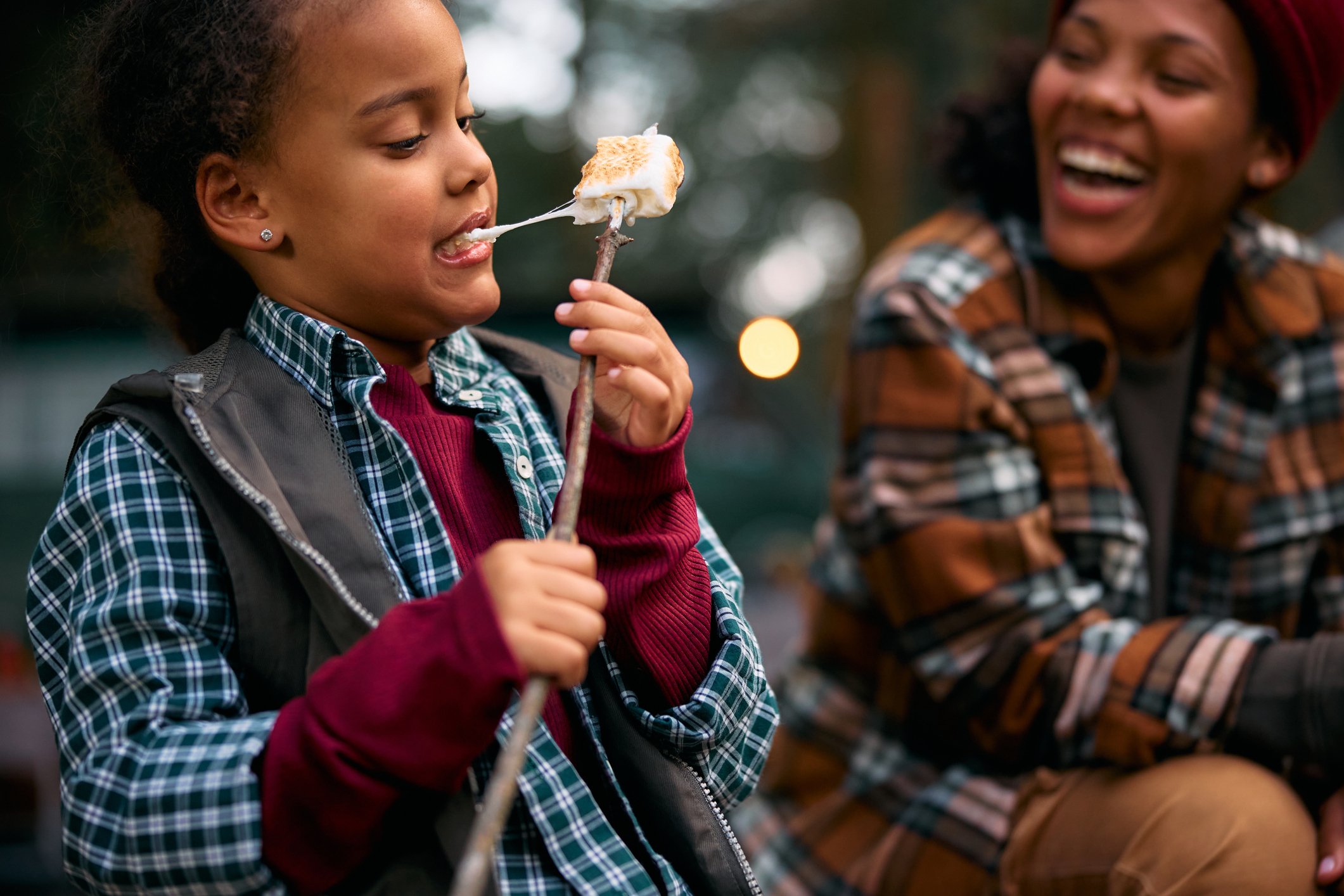 A young girl with earrings eating a marshmallow on a stick, smiling, with a woman in the background laughing.