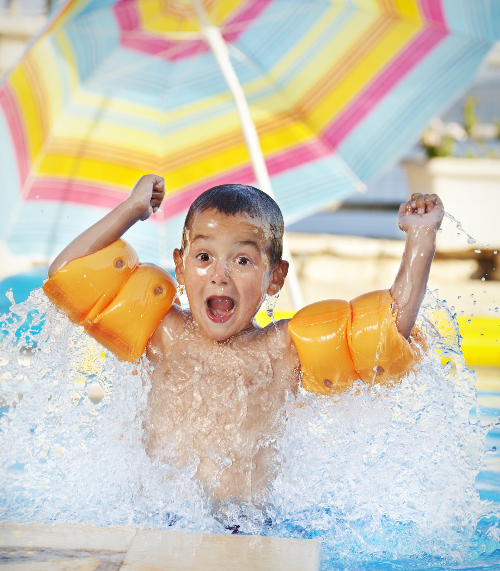 A young child laughing and splashing water in a swimming pool, wearing orange floaties and a colorful umbrella in the background.