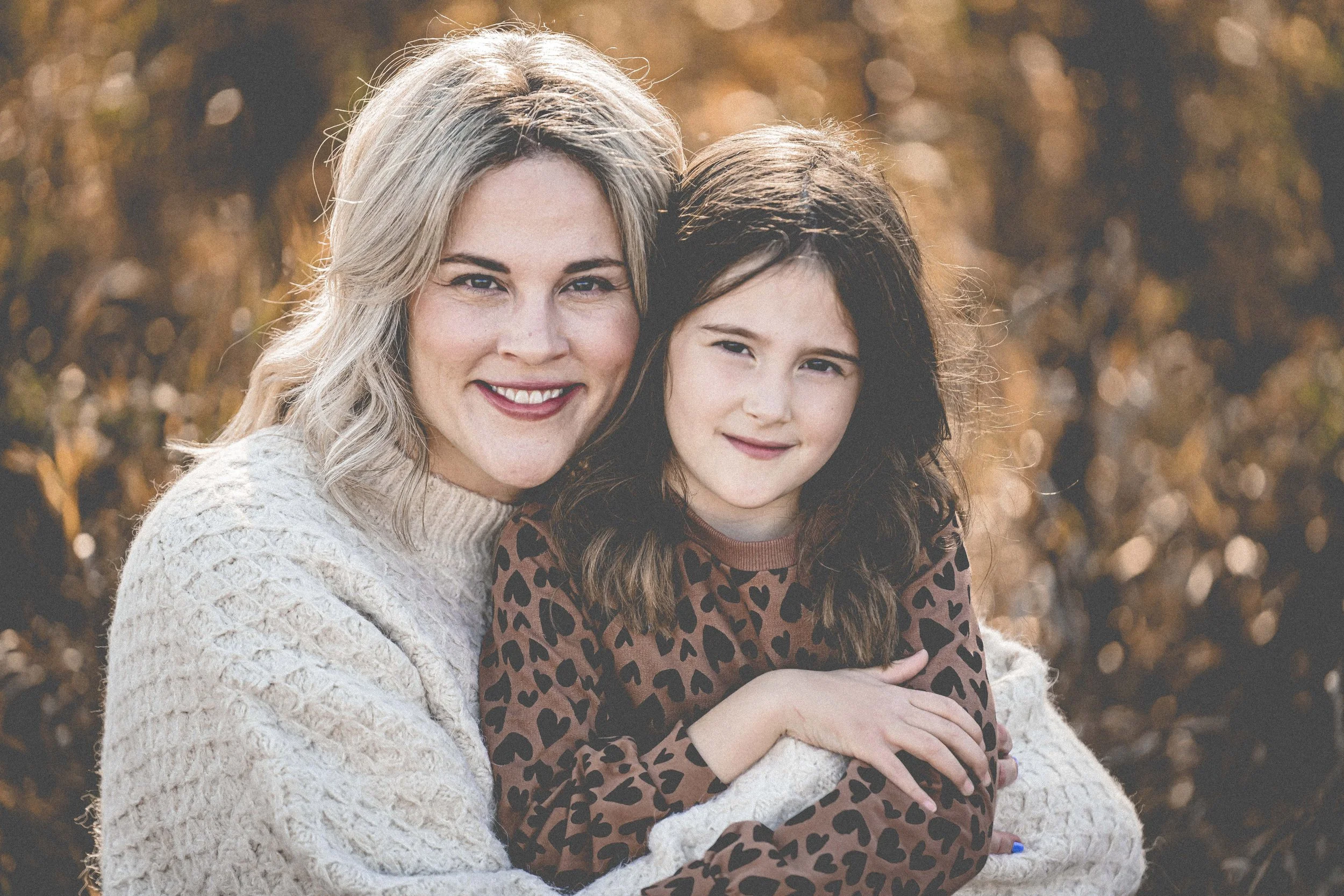 A woman with blonde hair and a young girl with dark, wavy hair, both smiling outdoors during autumn, with blurred fall foliage in the background.