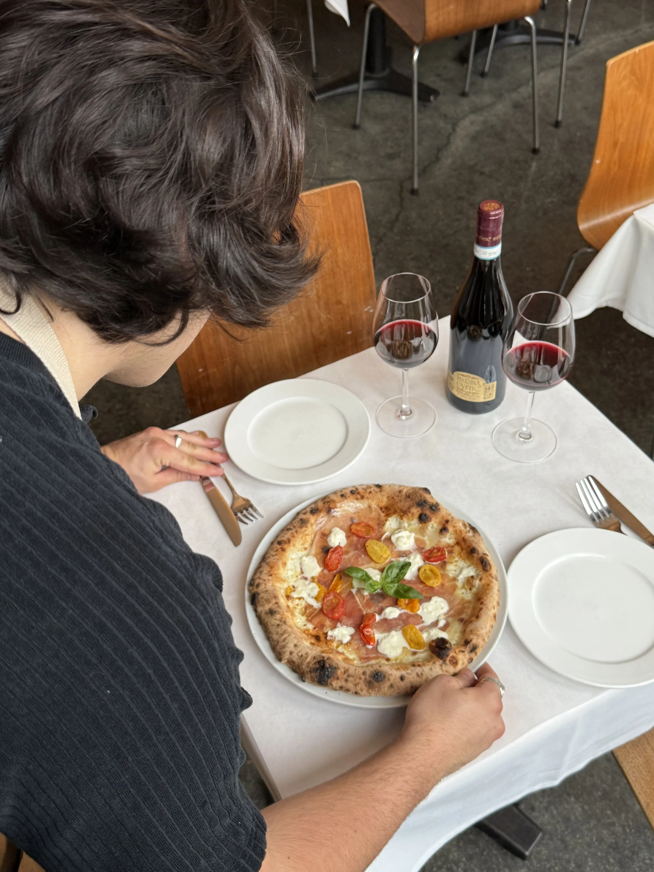 Person sitting at a table with a pizza, two glasses of red wine, a wine bottle, and empty plates in a restaurant.