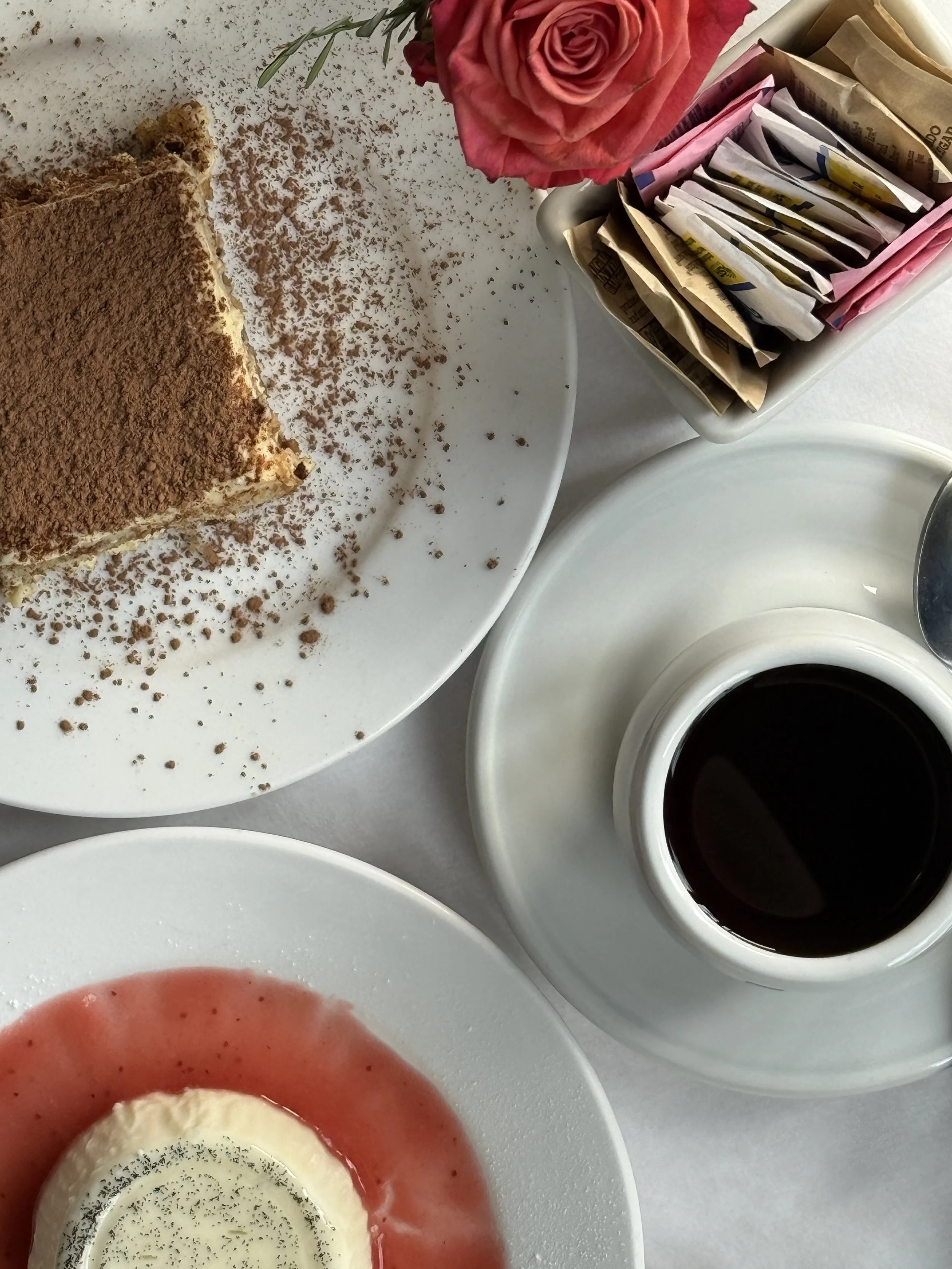 A white plate with slices of strawberry, whipped cream, and a sprinkle of black pepper or small seeds, surrounded by a bowl of black coffee, a container with sugar packets and sweetener, and a pink rose in a vase.