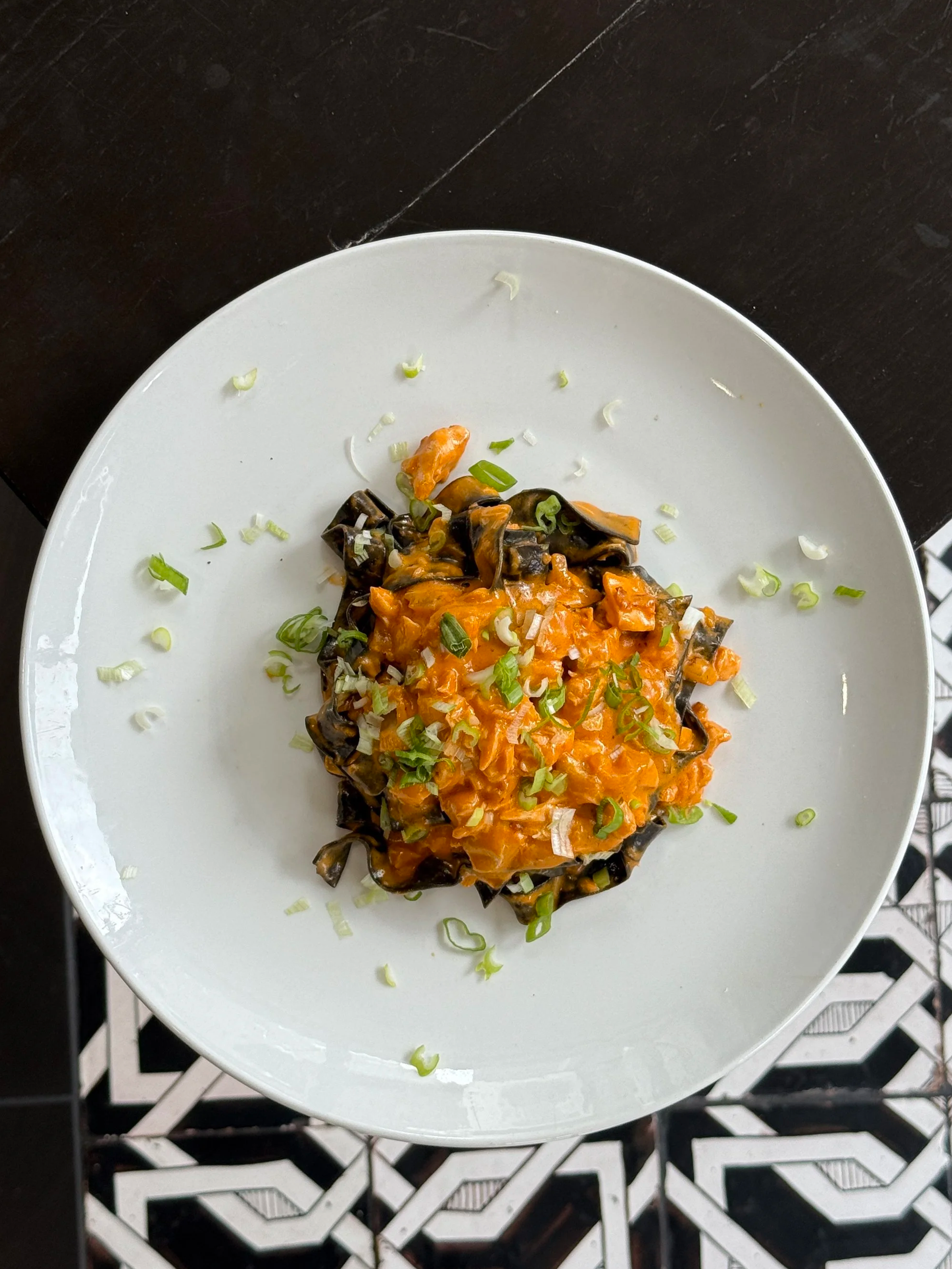A white plate with a serving of pasta topped with orange-colored tomato-based sauce, garnished with chopped green onions, placed on a black surface.