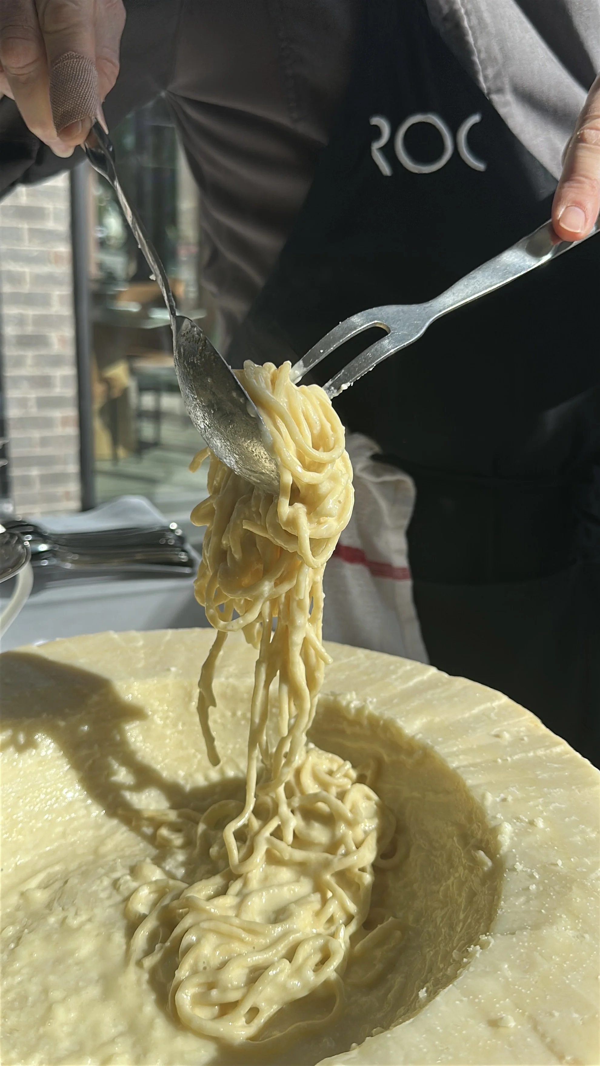 A person scoops cooked spaghetti noodles from a large wheel of Parmesan cheese.