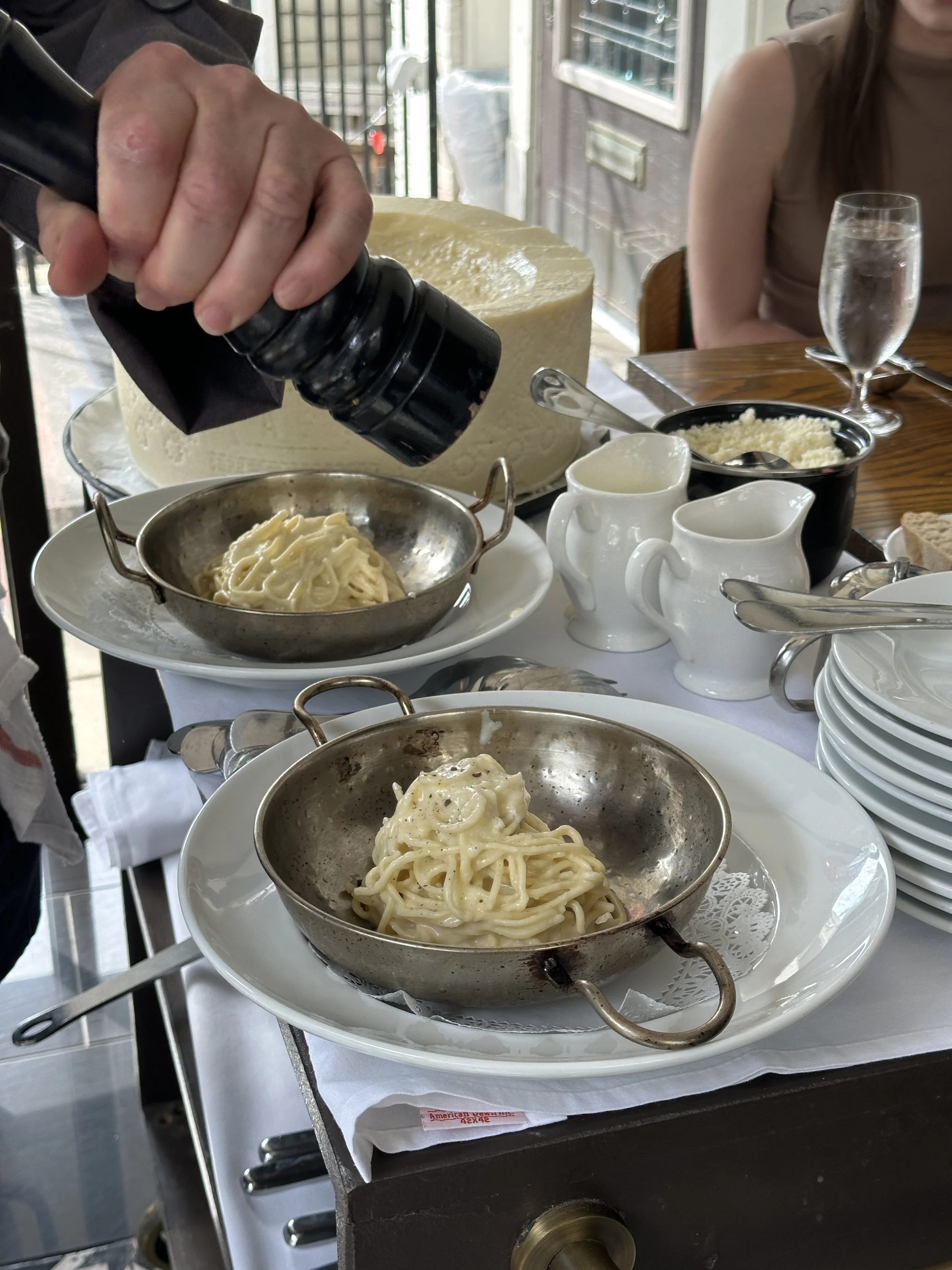 Hand sprinkling grated cheese onto plates of spaghetti pasta at a restaurant table with water glasses and small pitchers.