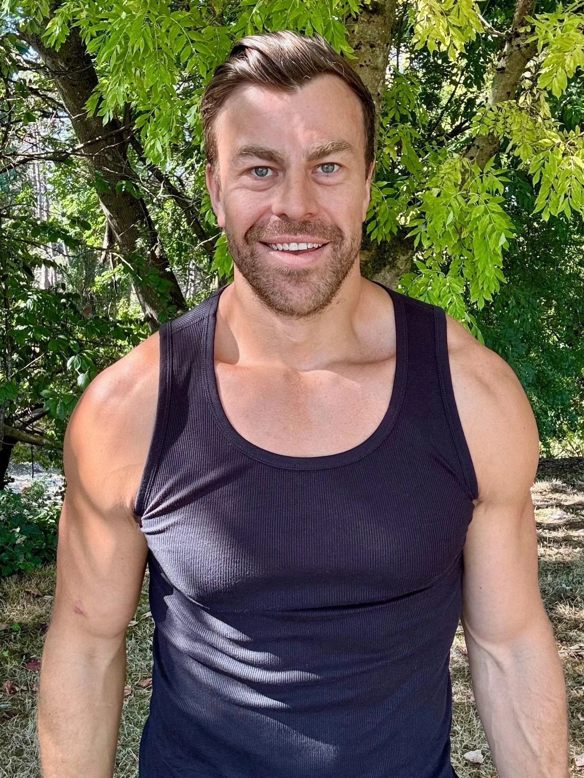 A smiling man with short brown hair and blue eyes, wearing a black tank top, standing outdoors in front of green leafy trees.