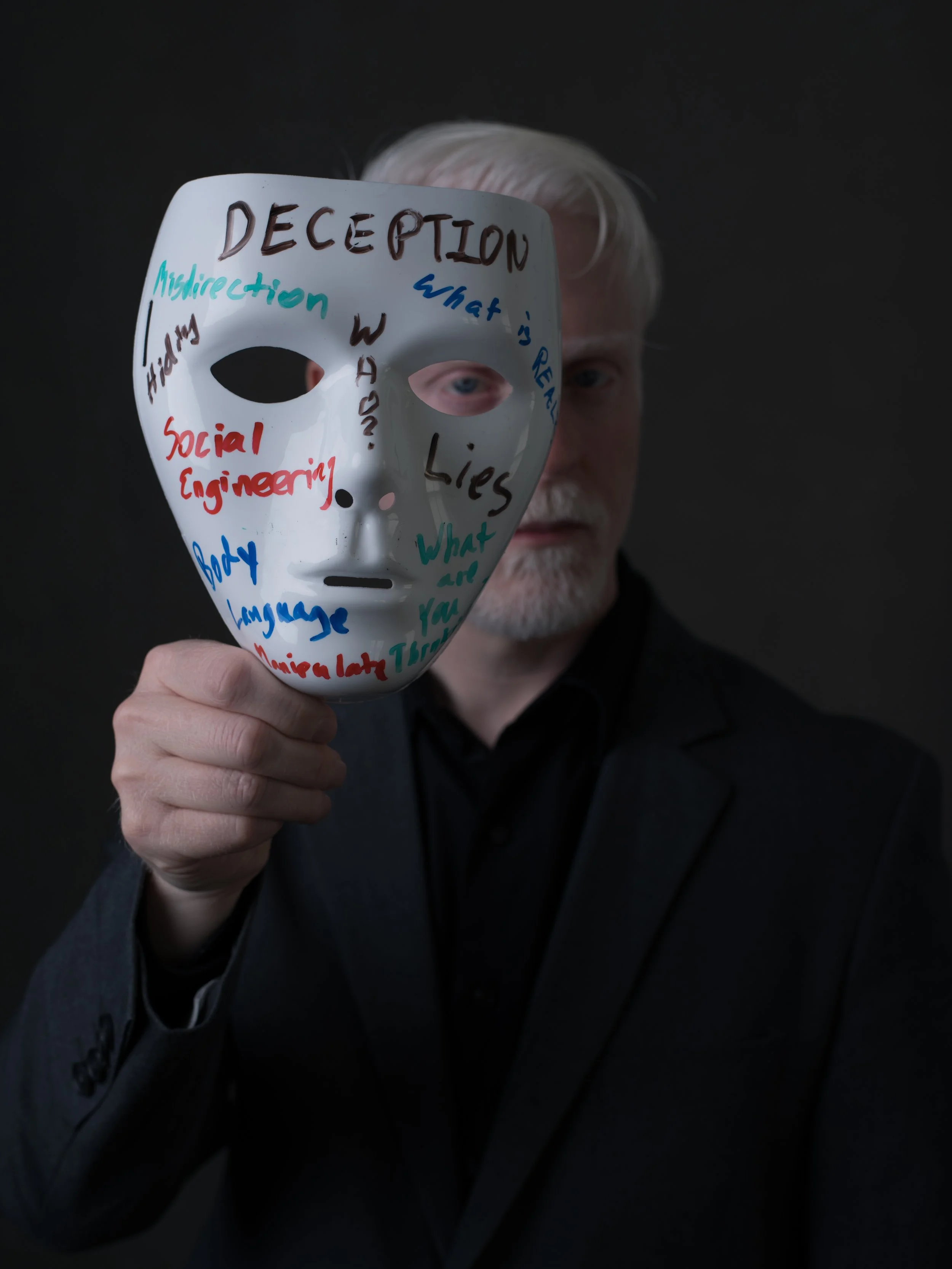 Perry Carpenter holding a mask with colorful words written on it, including 'DECEPTION', 'Misleading', 'Lies', 'Social engineering', and 'What is real?'.