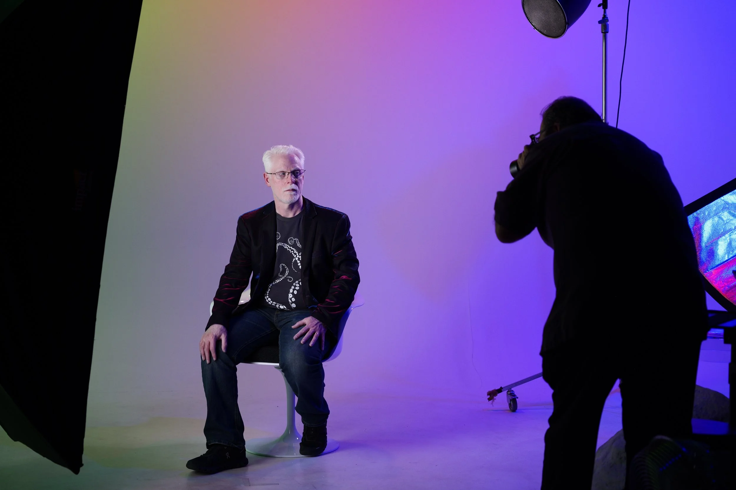 Behind the scenes shot of Perry Carpenter sitting on a stool during a photo shoot, with a photographer taking pictures against a colorful gradient background.