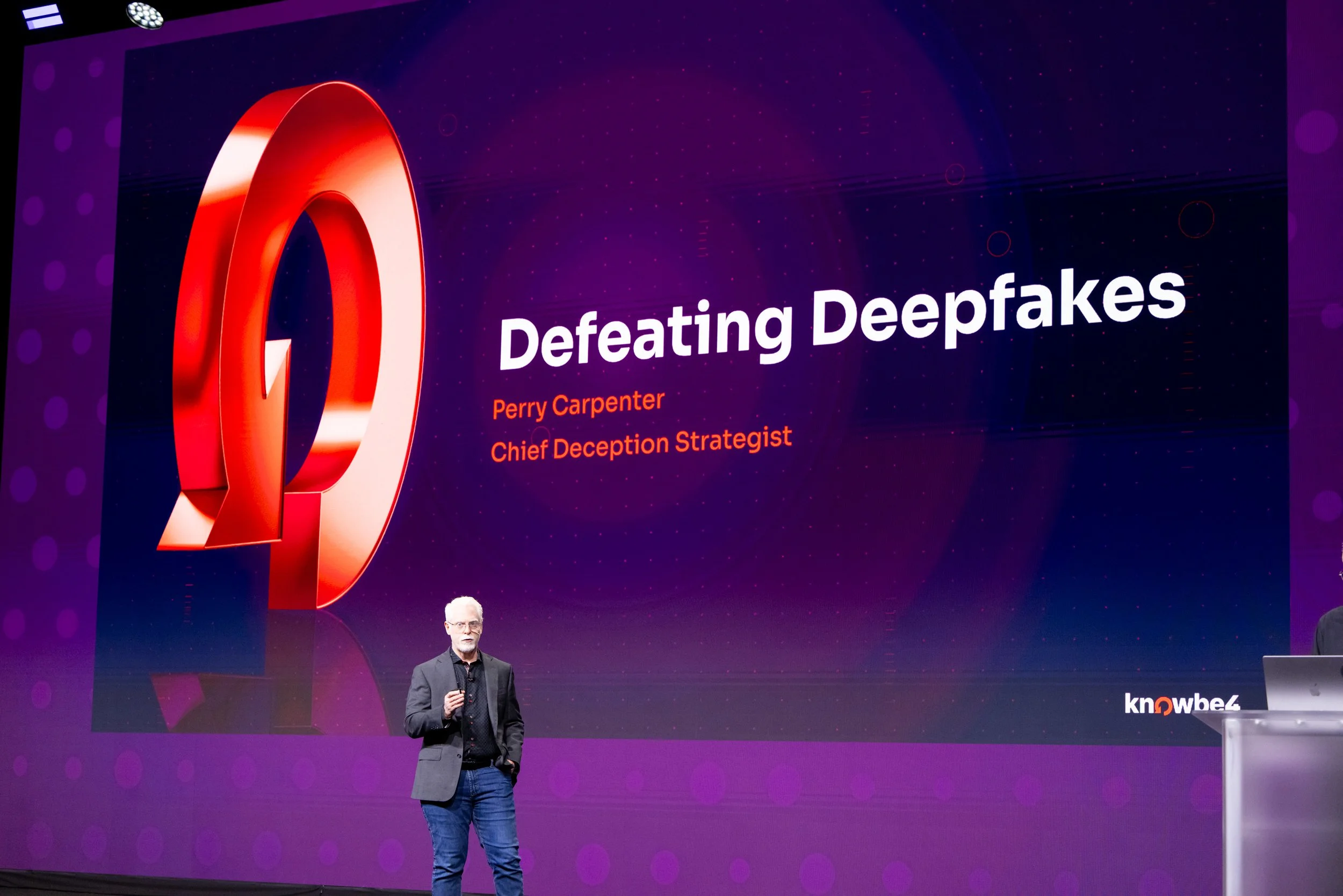 A man is giving a presentation on stage with a large screen behind him. The screen displays a headline 'Defeating Deepfakes' with the name Perry Carpenter and his title 'Chief Deception Strategist.' The stage background is purple with dot patterns, and the man is dressed in a gray blazer, black shirt, and jeans.