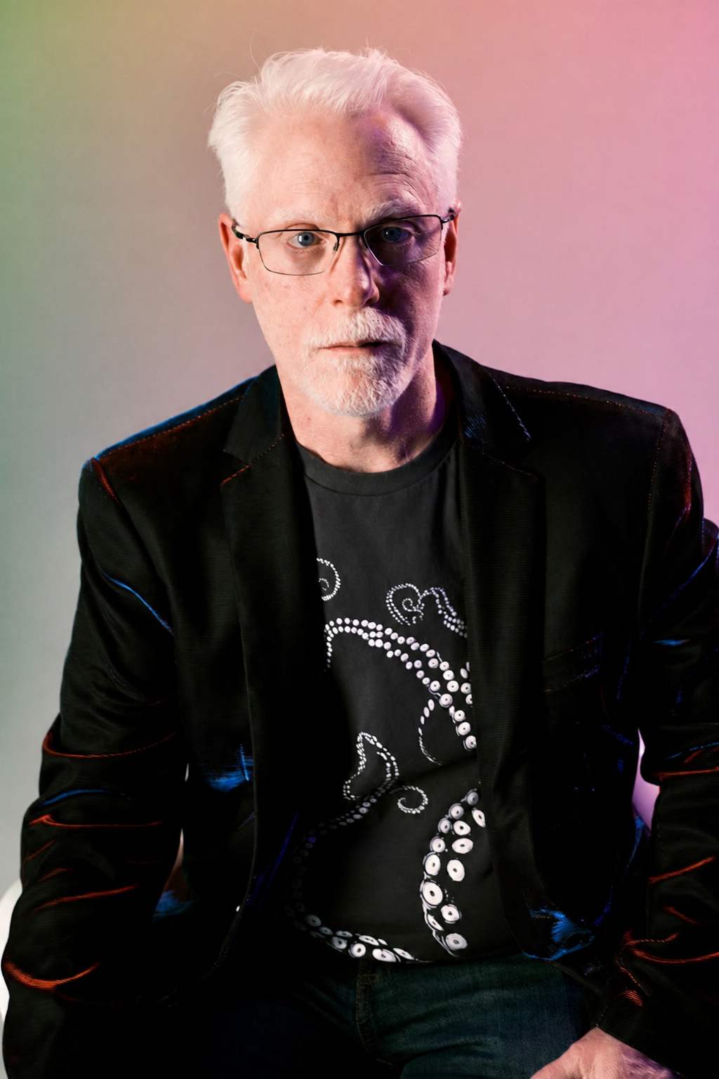Perry Carpenter wearing a jacket over a T-shirt with a white octopus design, sitting against a gradient background with soft pink, purple, and green hues.