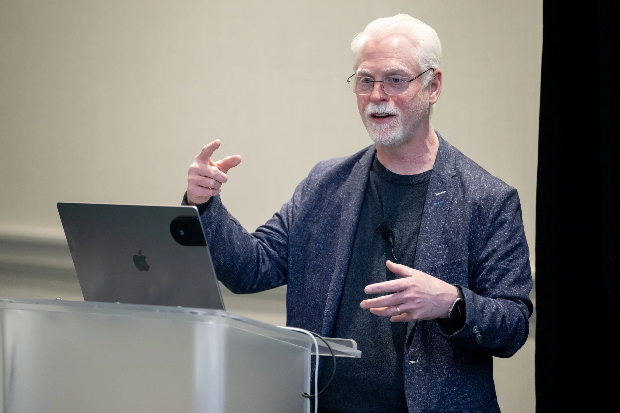 A man with white hair and glasses is standing at a podium, wearing a dark blazer and speaking. He is pointing with his right hand and appears to be engaged in a presentation or speech, with a laptop in front of him.