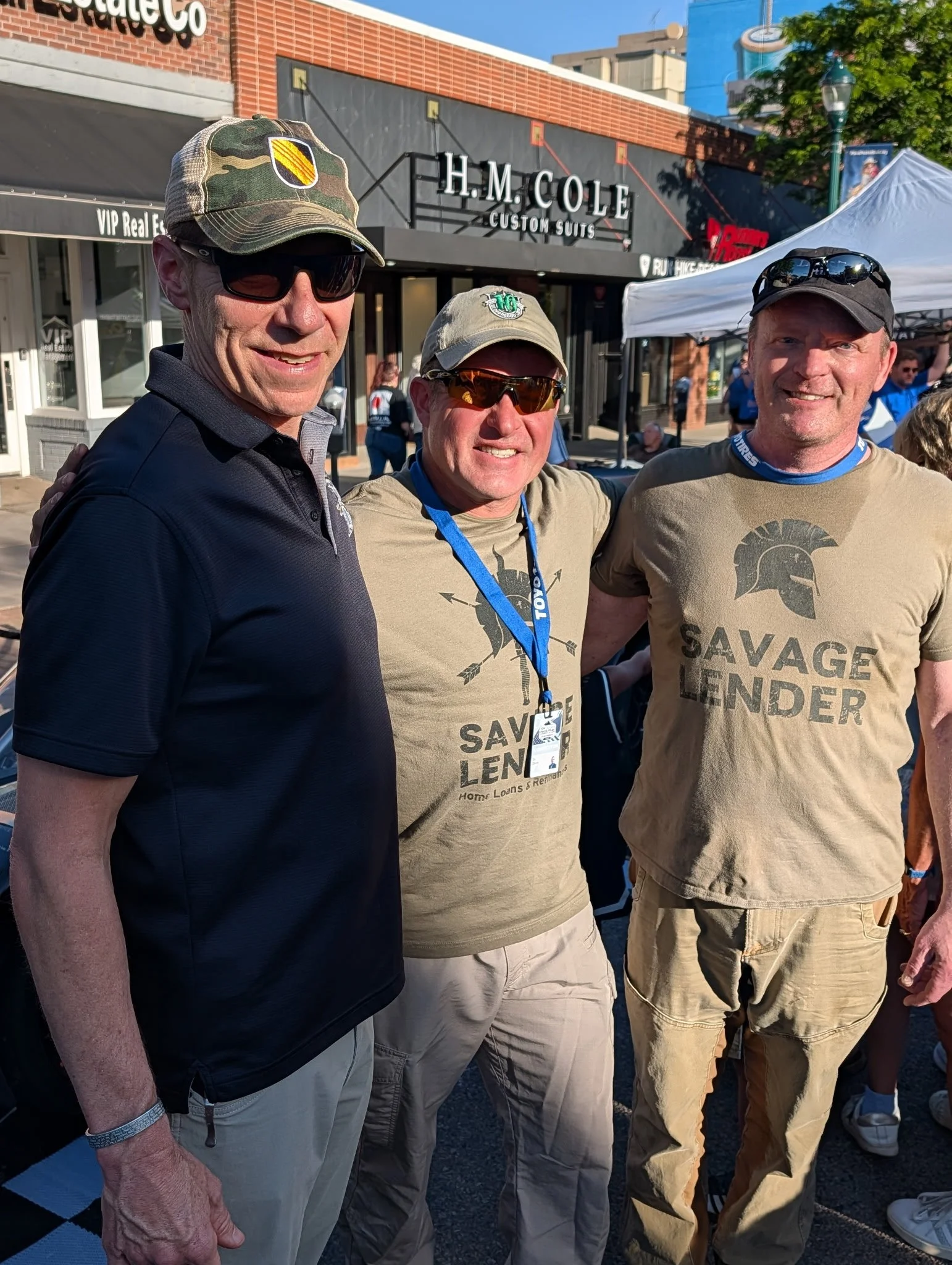Three men standing together outdoors at an event, with shops and a white tent in the background. They are smiling and are dressed casually, wearing caps, sunglasses, and t-shirts, some with printed text and graphics.