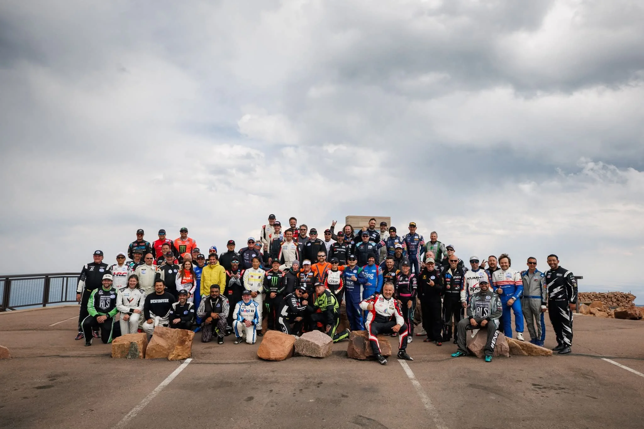 Group of race car drivers and team members gathered outdoors on a cloudy day, standing behind large rocks, at a racing event or track.