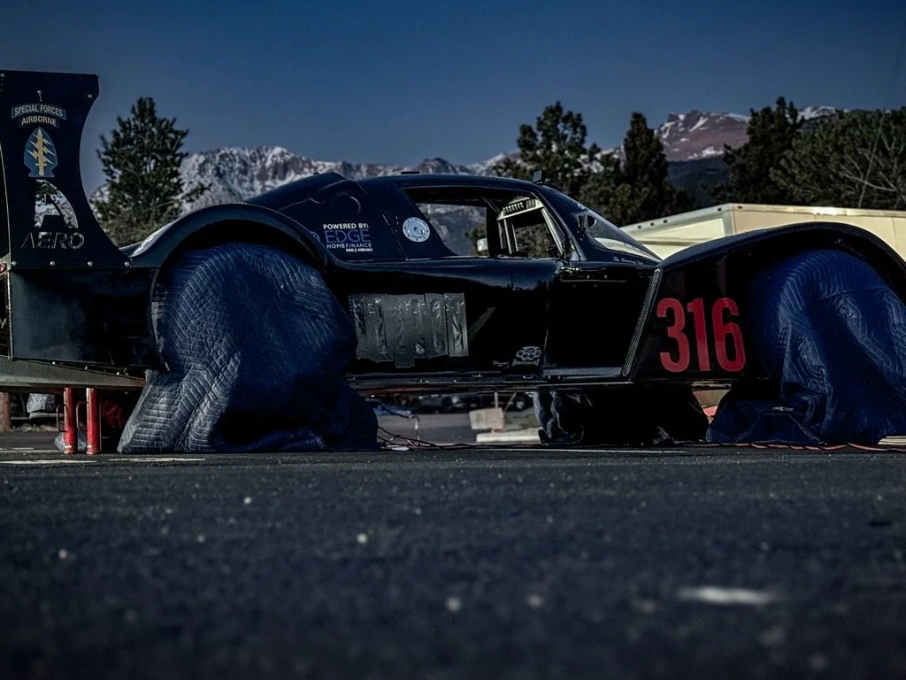 A black racing car with the number 316, parked outdoors on a paved surface with mountainous terrain and trees in the background.
