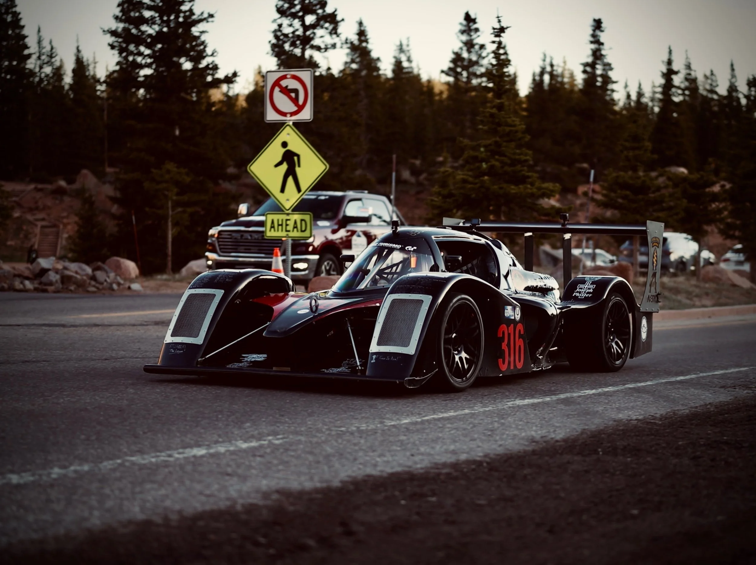 A race car on a road with trees in the background, with a yellow pedestrian crossing sign and a no left turn sign.