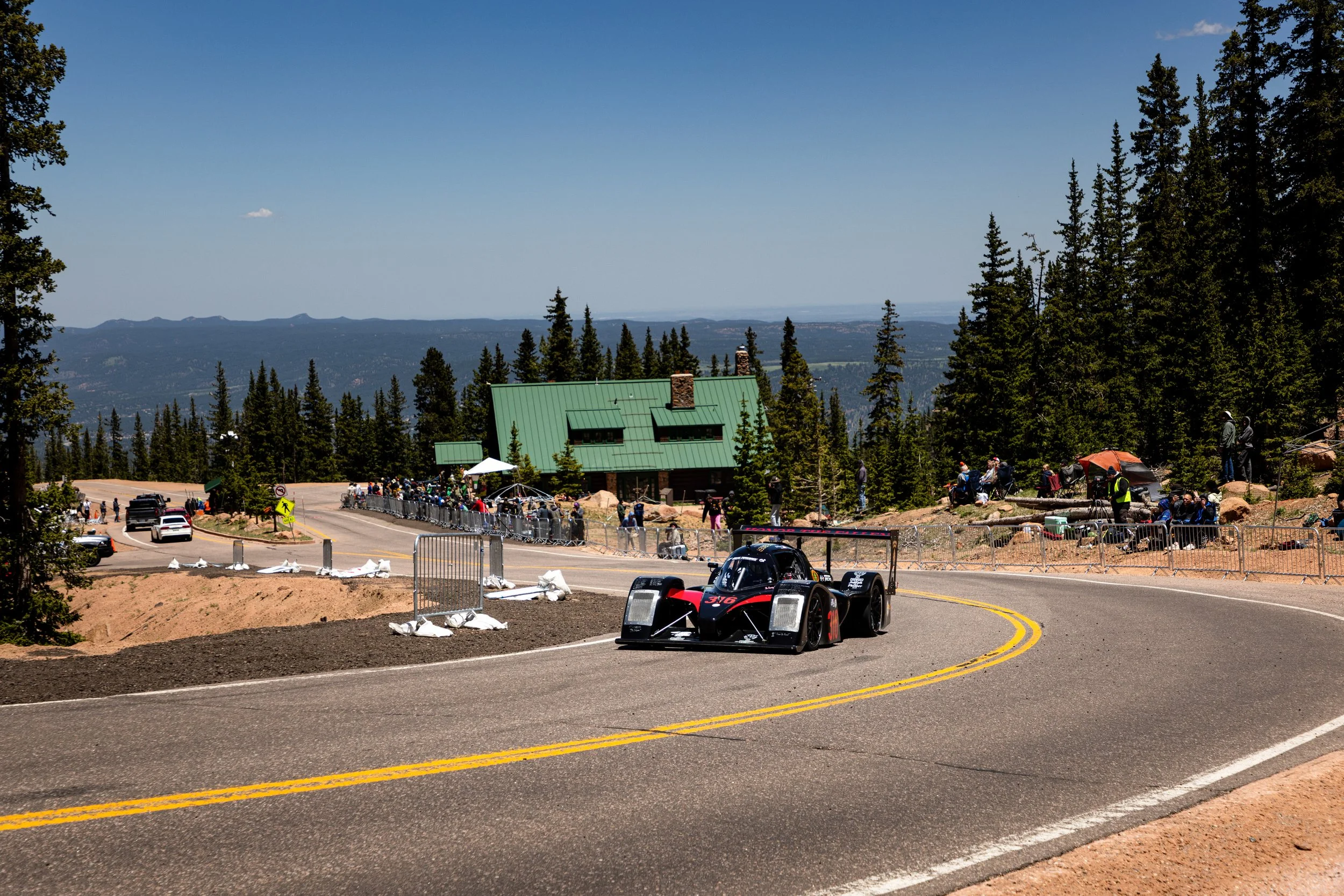 A race car on a winding mountain road with spectators and a green-roofed building in the background, surrounded by tall pine trees.