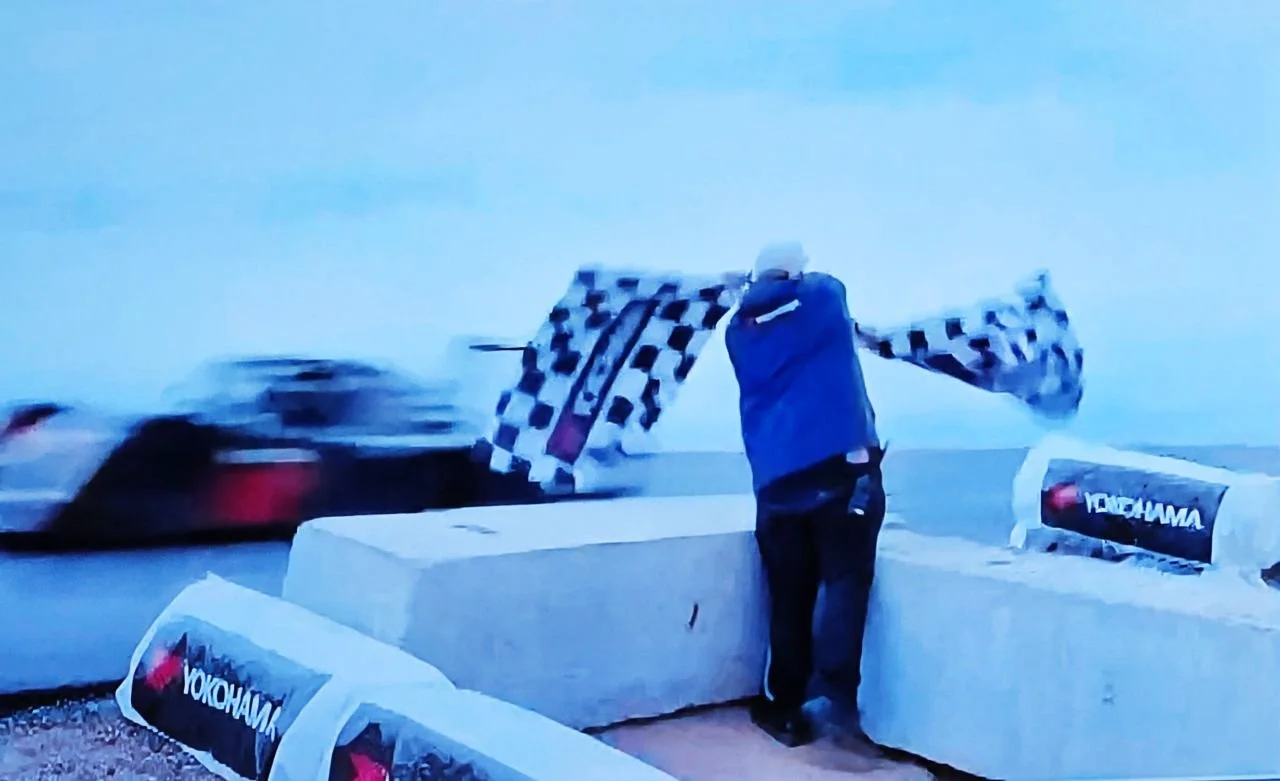 Person holding checkered flag at race finish line with racing car blurred in motion in the background.