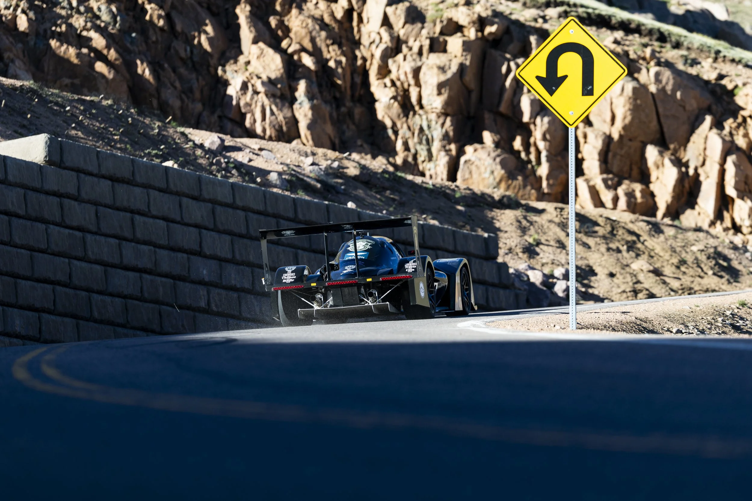 Close-up view of a black race car on a mountain road behind a roadblock, with a yellow U-turn warning sign in the background.