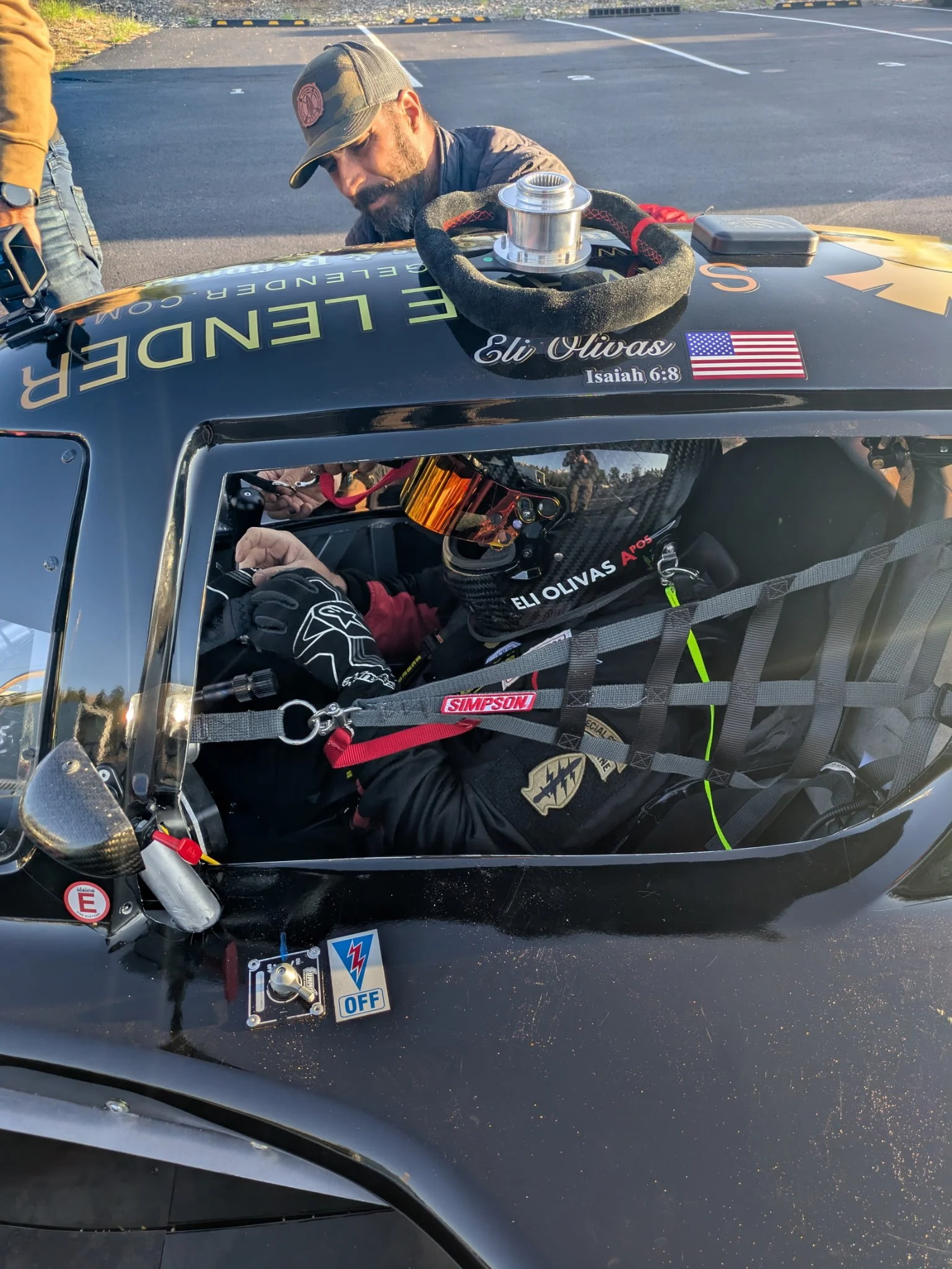 A man working on a race car in a parking lot, wearing a cap and gloves, with various tools and equipment visible inside the car.